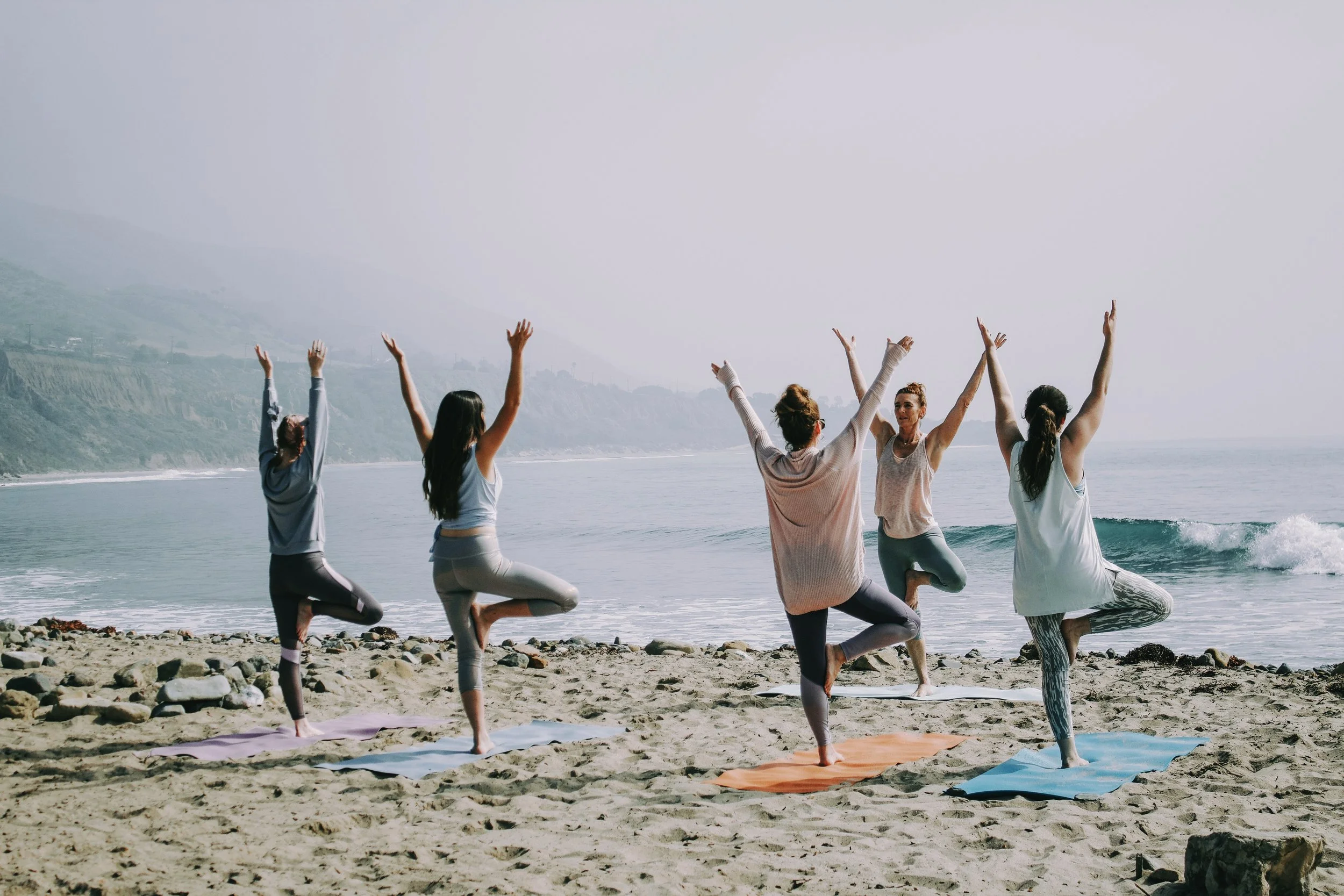 A group of five women practicing yoga on the beach during daytime, standing on one leg with the other foot resting on the inner thigh, arms raised overhead, facing the ocean.