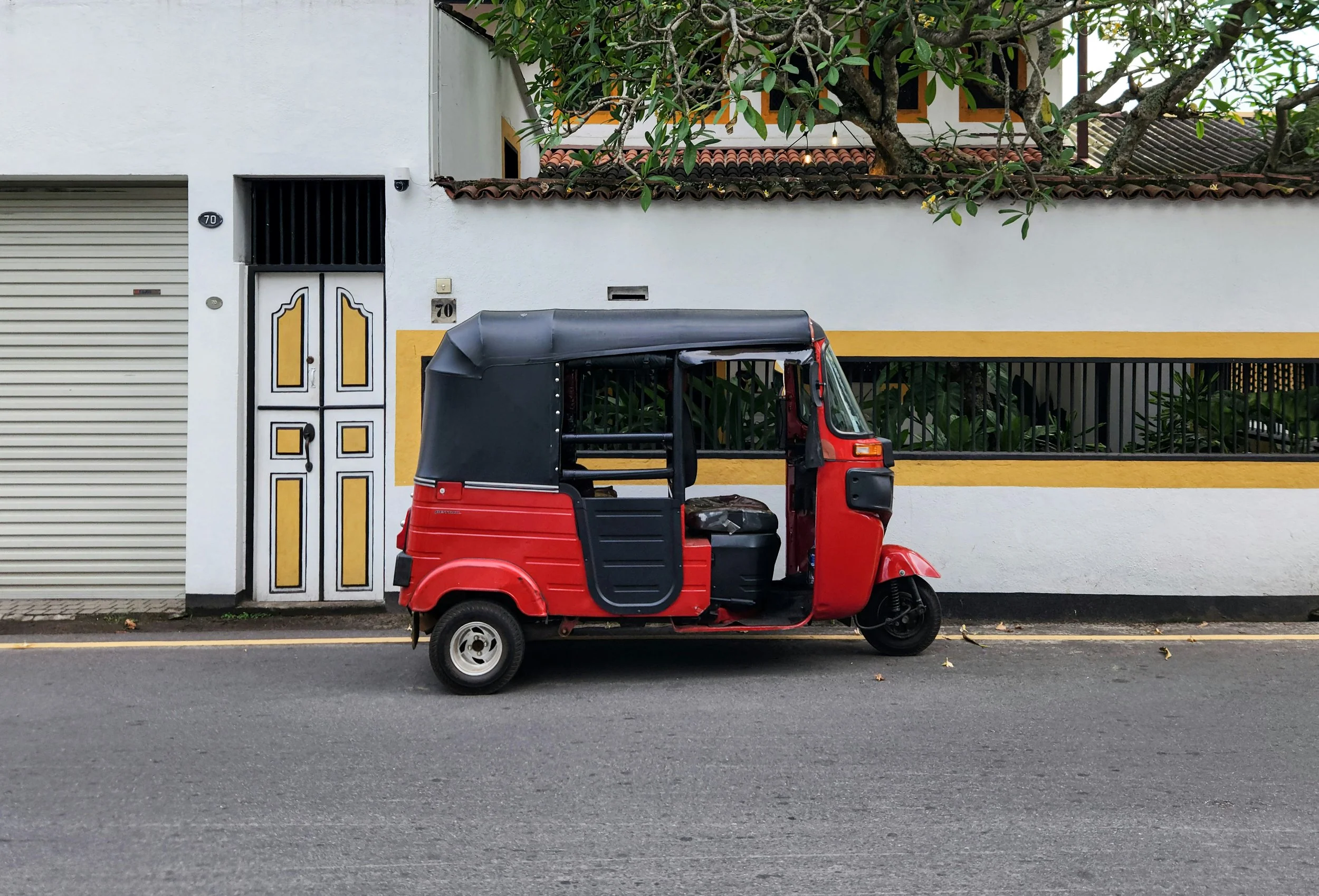 A red and black tuk-tuk parked on the street in front of a white wall with yellow accents, a gate, and a tree with green leaves.