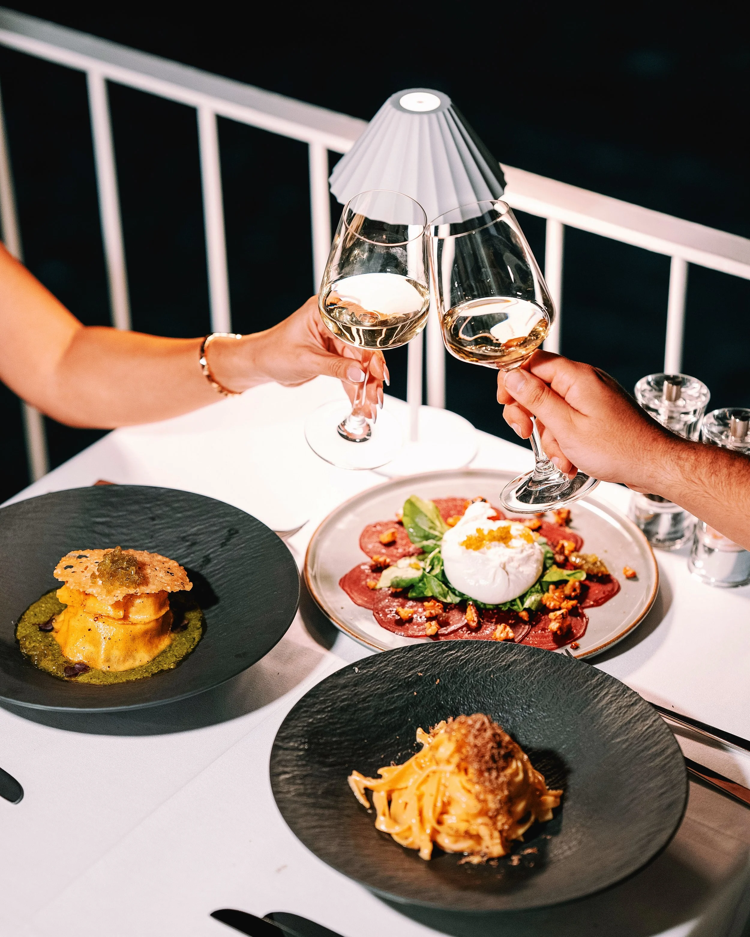 Two people clinking glasses of white wine over a table with assorted gourmet dishes, including a dessert, with a black slate plate, a salad, and a lamp in the background.
