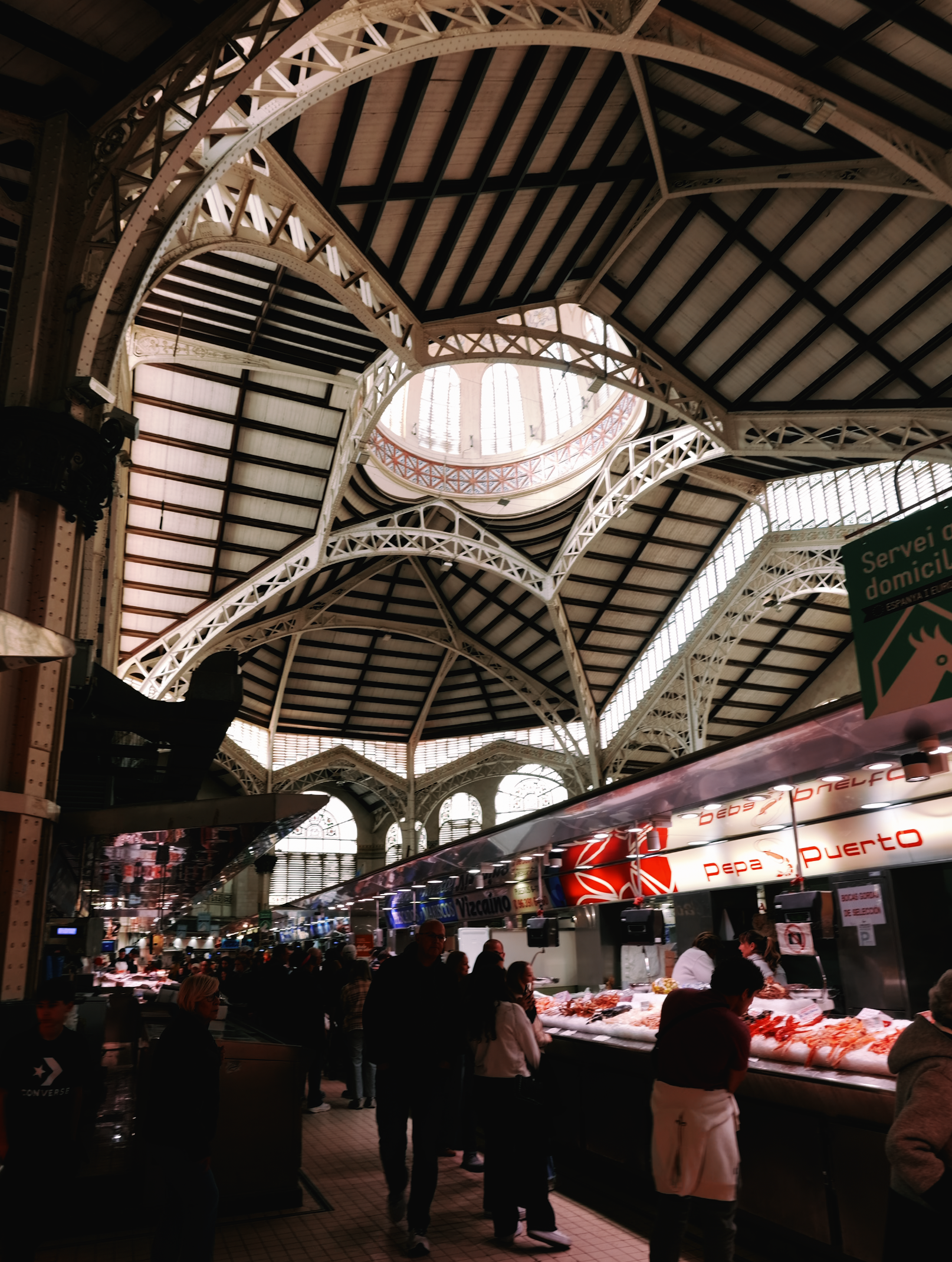 Inside a busy indoor market with a high arched ceiling supported by intricate metal framework, customers browse seafood displayed on counters, with signs and advertisements overhead.