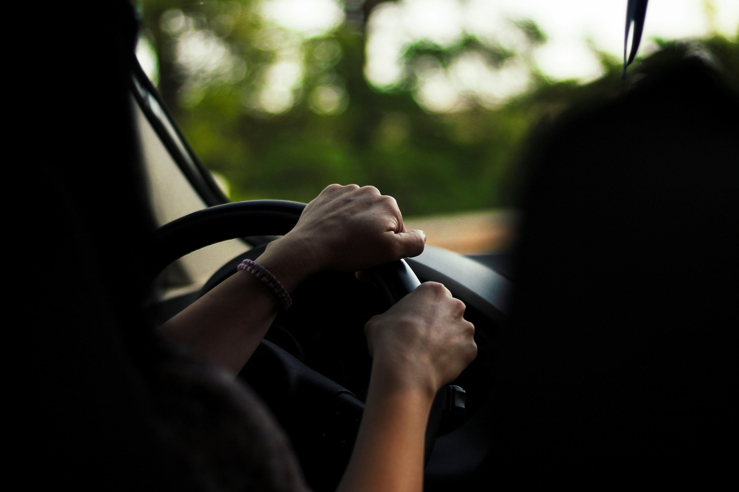 A person’s hand gripping a steering wheel while driving, with greenery visible through the windshield.