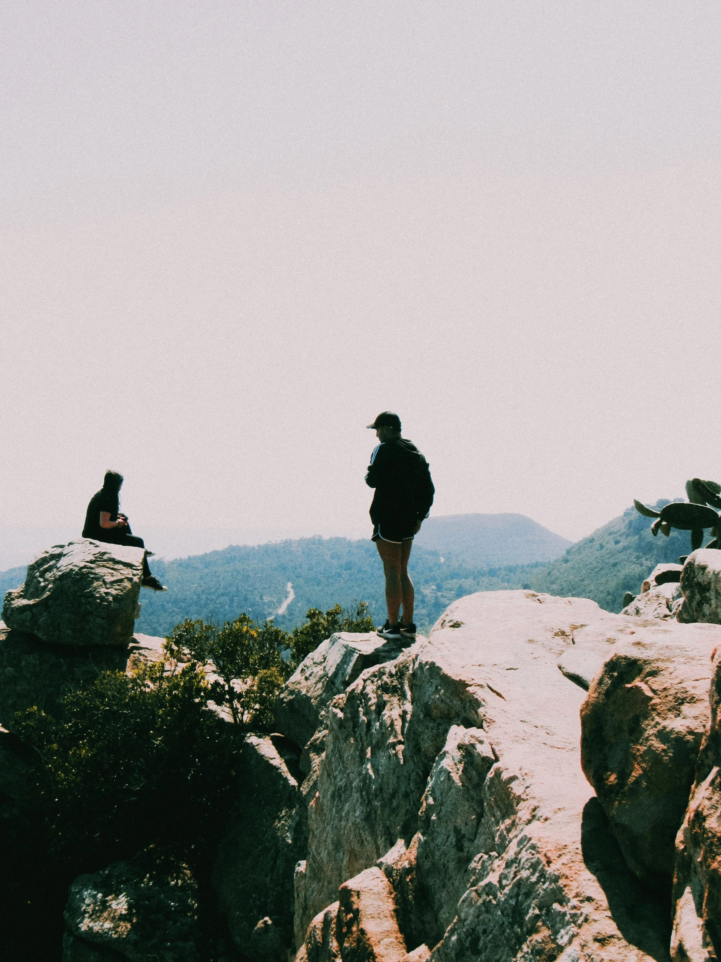 People hiking on rocky terrain overlooking a mountainous landscape with trees, distant mountains, and sparse clouds in the sky.