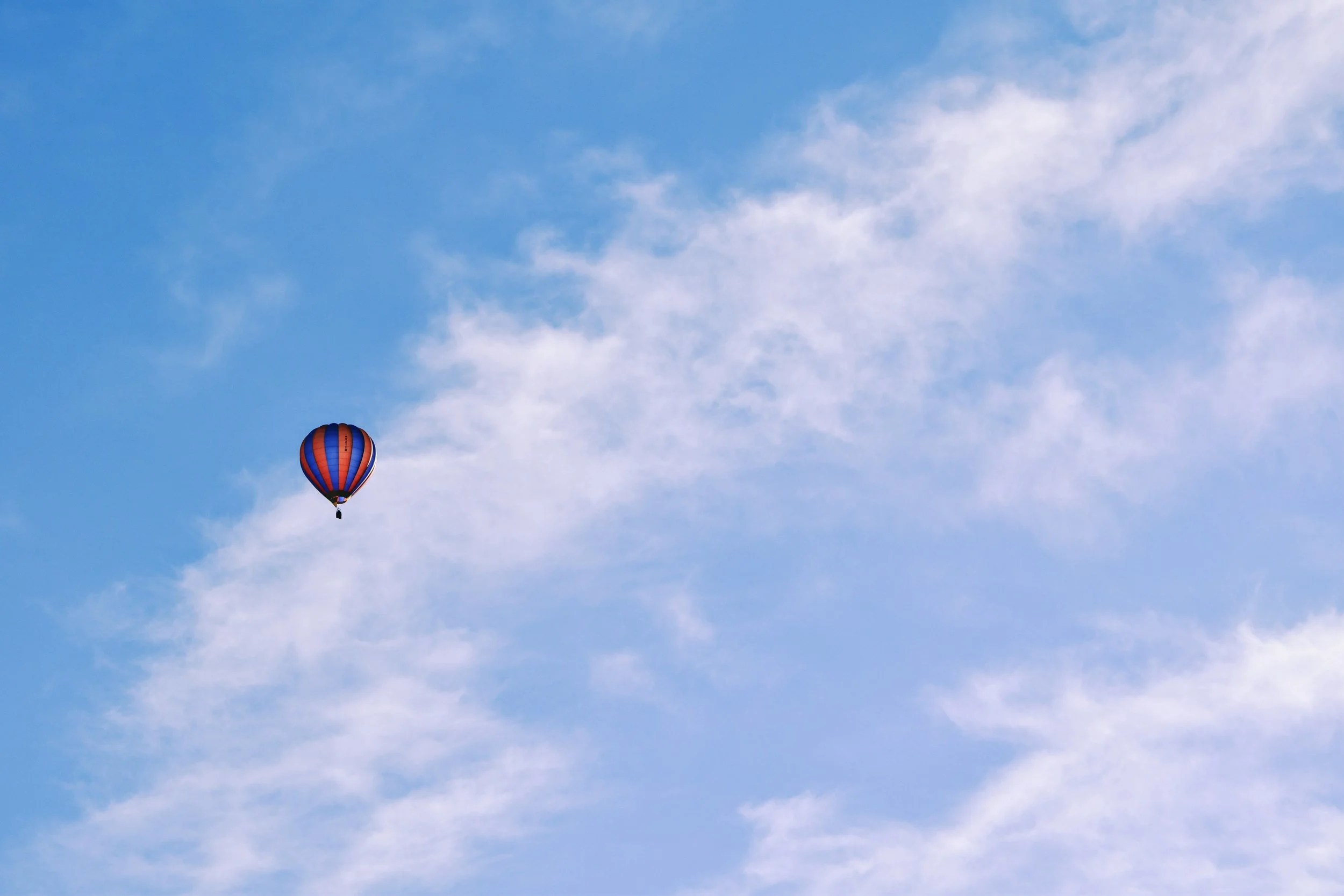 A hot air balloon with red, blue, and black vertical stripes floating in a sky with light clouds.
