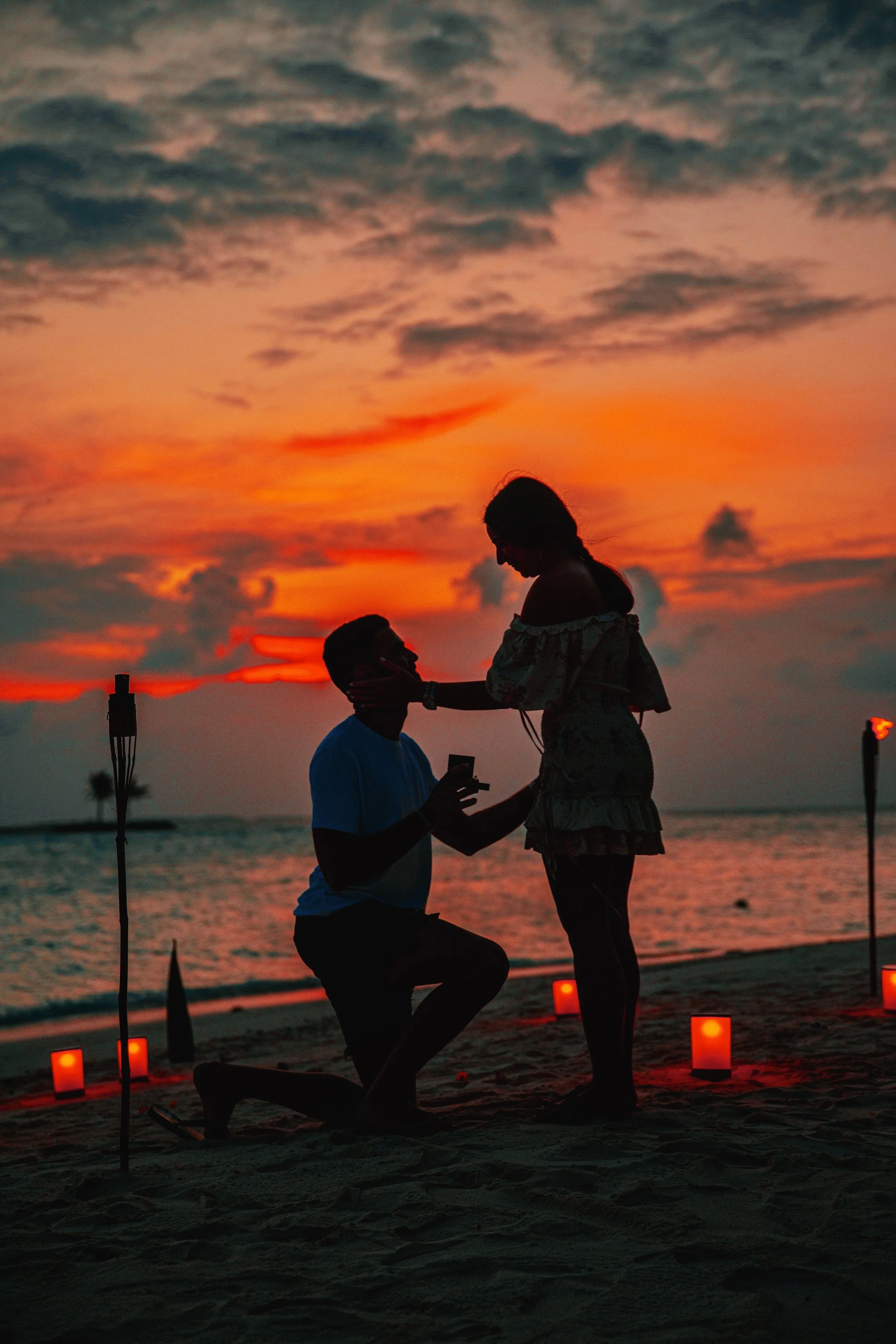 A couple exchanging rings during a sunset on a beach, with decorative lanterns and tiki torches around them.