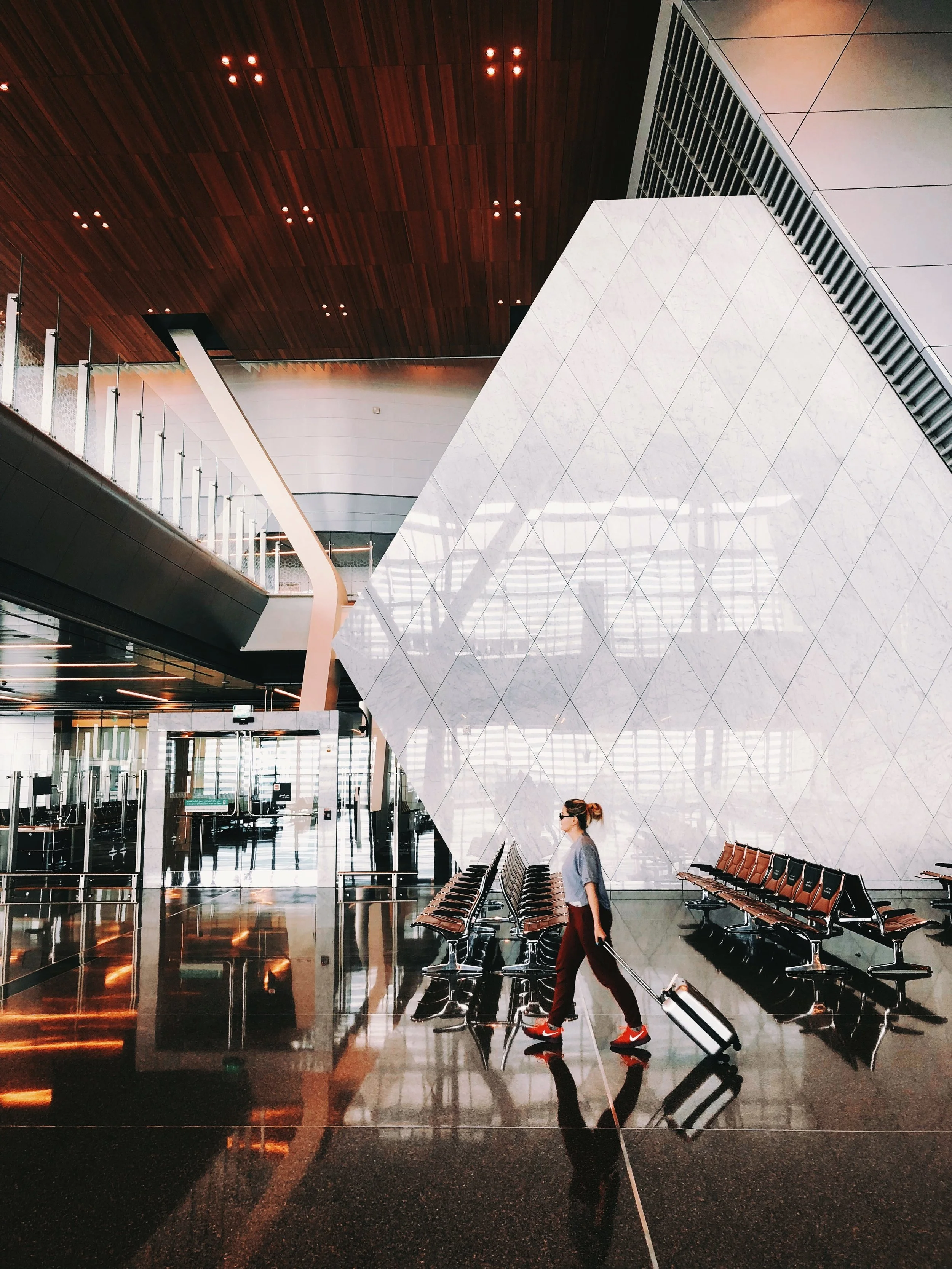 An airport terminal with modern design, wooden ceiling, white geometric wall, and rows of seating. A woman is walking with a rolling suitcase.