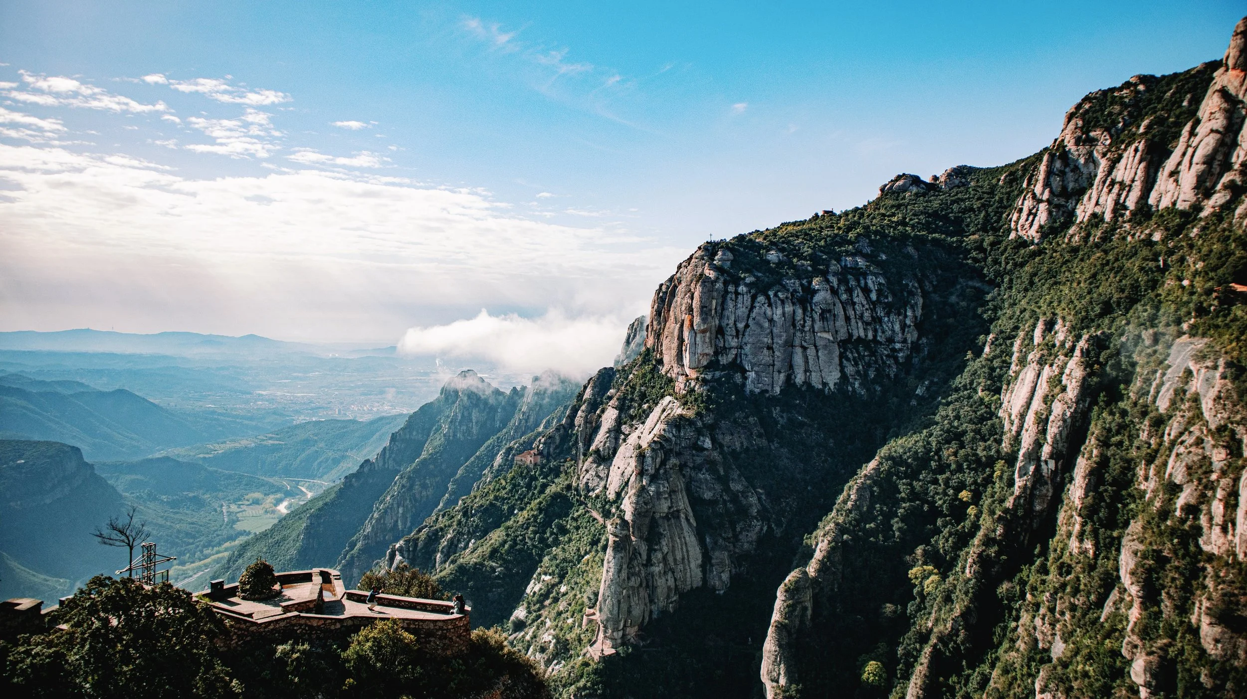 Mountain landscape with steep rocky cliffs covered in green vegetation, distant hills, and valleys under a partly cloudy sky.