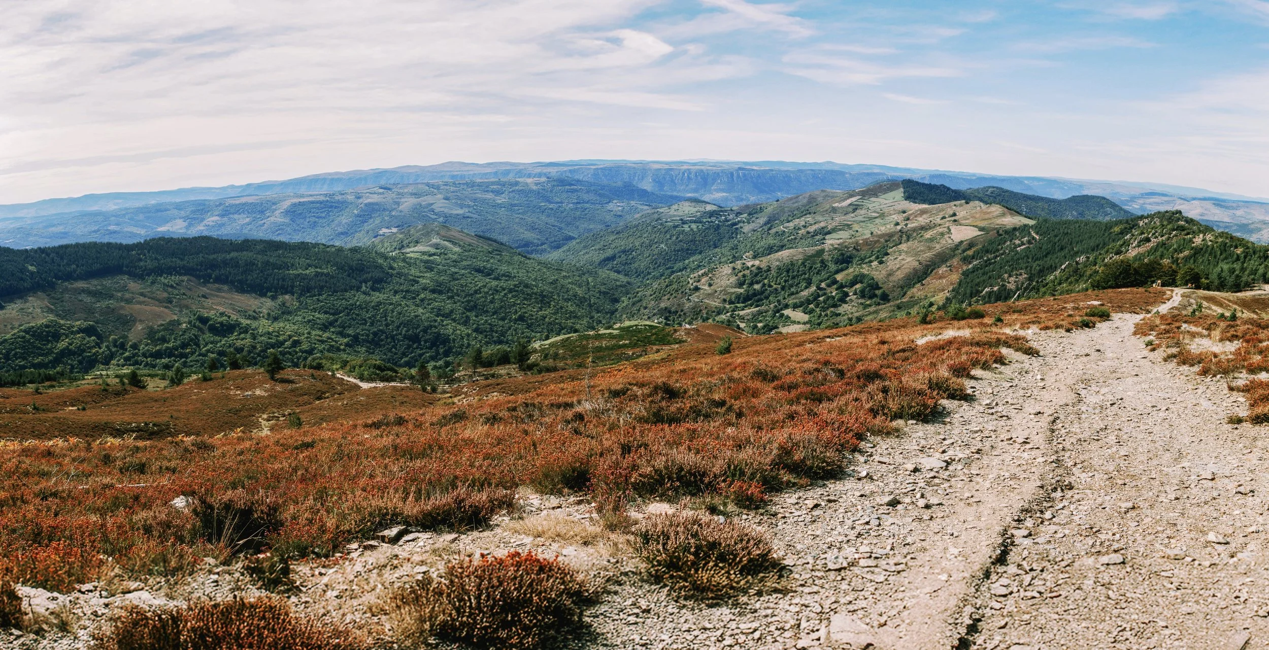 A mountain landscape with a dirt trail winding through reddish-brown and green vegetation, rolling hills, and distant blue mountains under a partly cloudy sky.