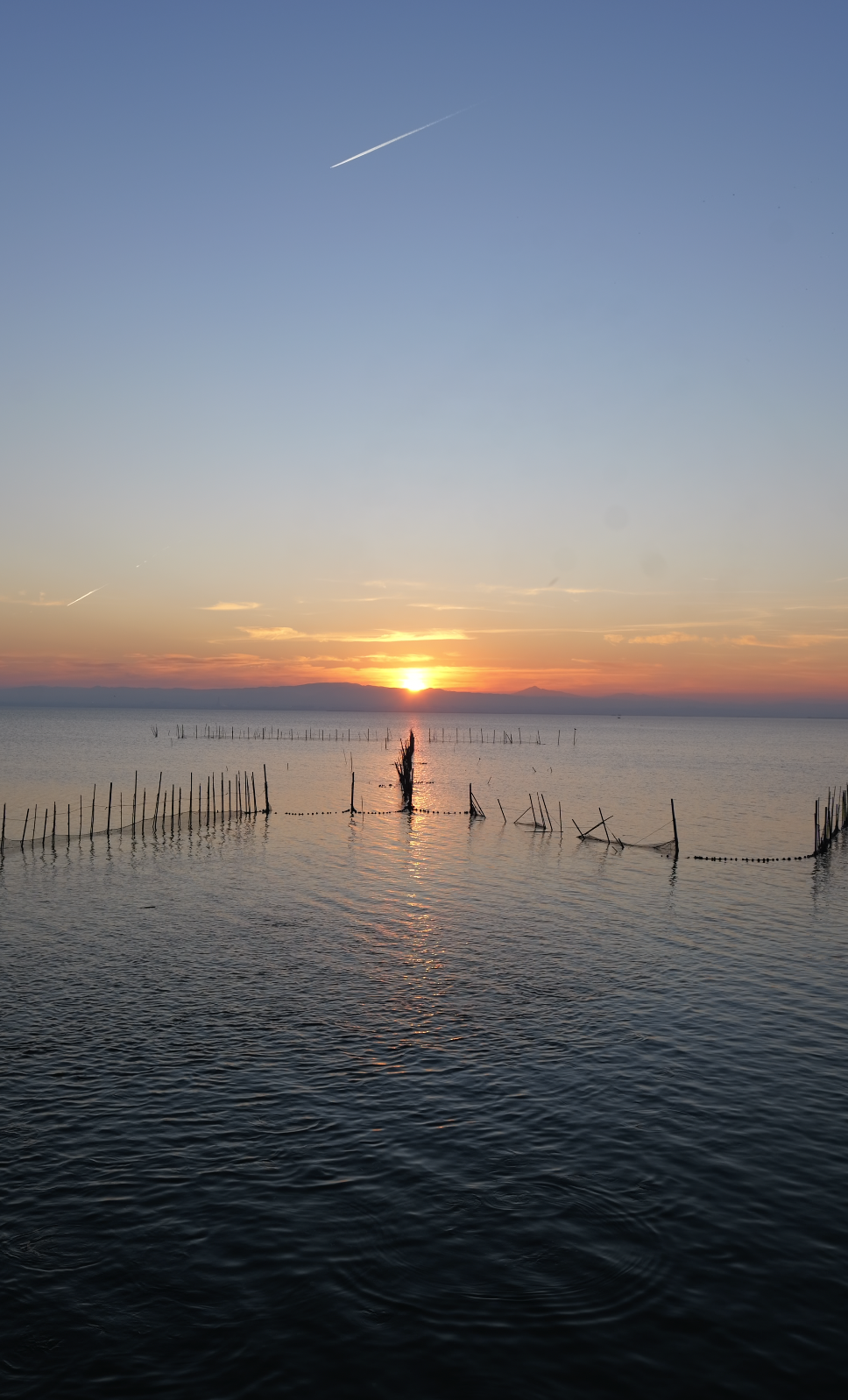 Sunset over a calm body of water with a partly cloudy sky and a contrail in the distance.