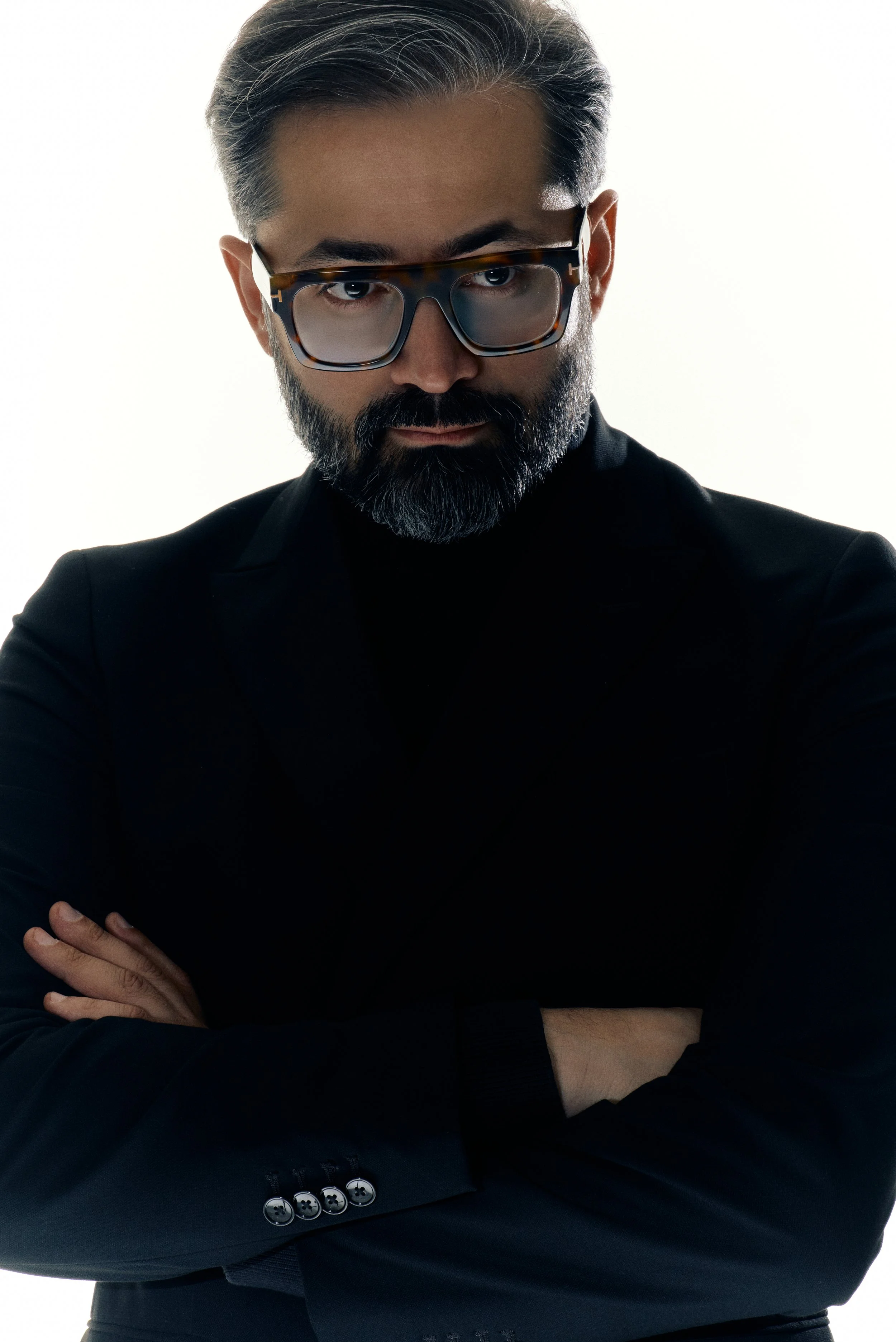 A man with gray-tinted hair and a beard, wearing dark sunglasses and a black suit jacket with buttons, with his arms crossed and a serious expression.