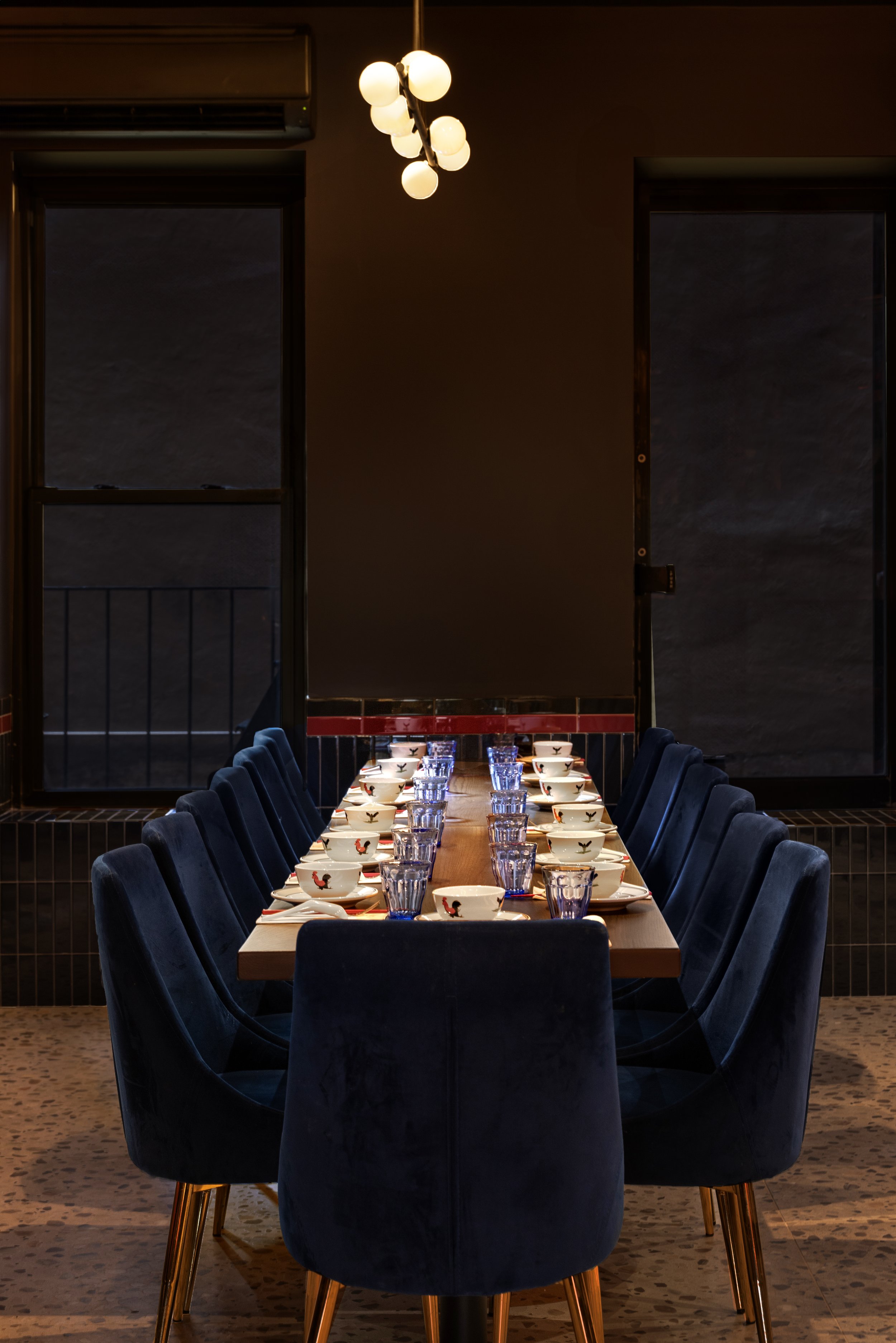 A long dining table set with bowls and glasses in a dimly lit restaurant or dining room with dark walls, window, and modern lighting fixture.