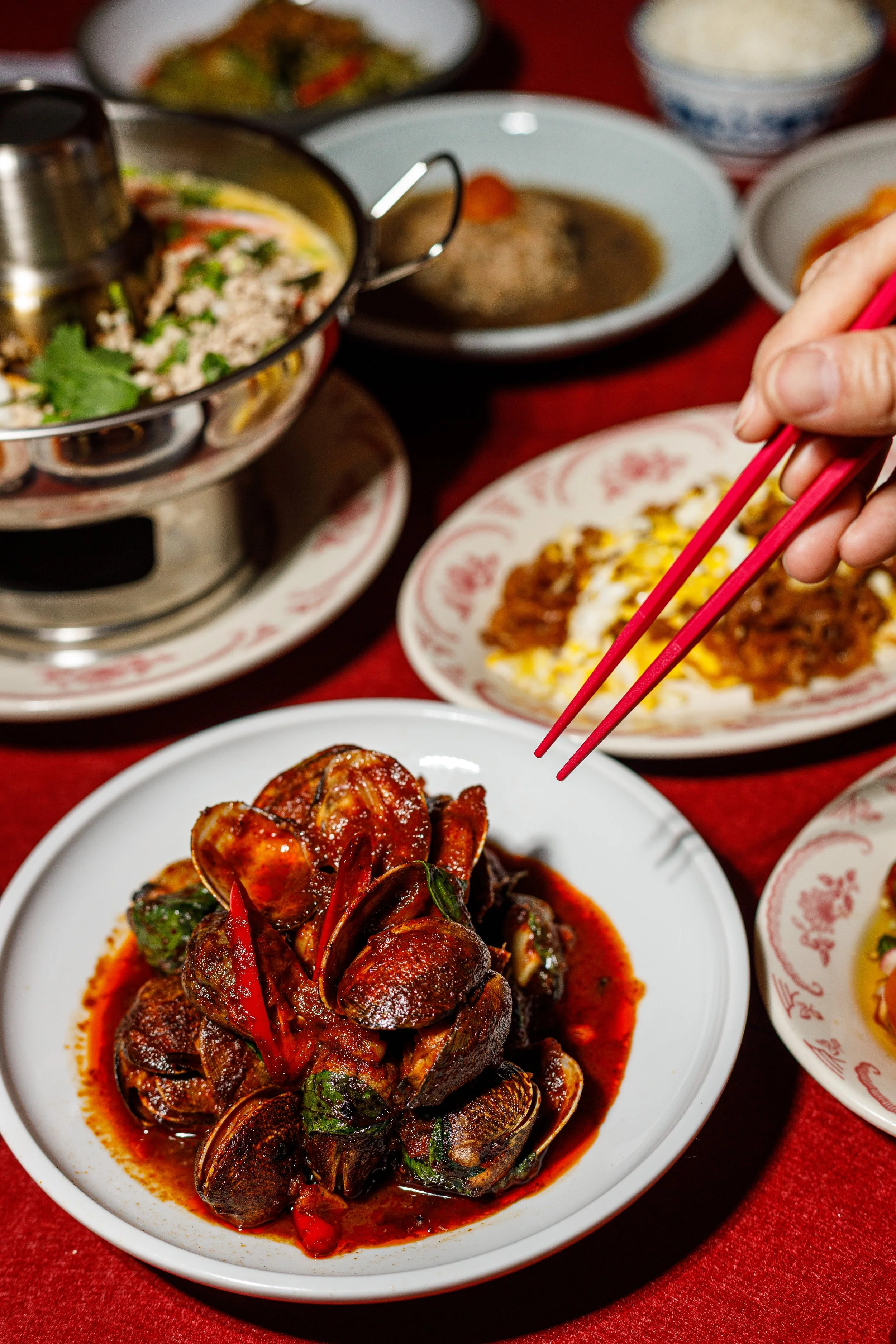 A Chinese seafood dish with mussels and red peppers in a spicy sauce on a white plate, surrounded by various Chinese dishes and bowls of rice on a red tablecloth.