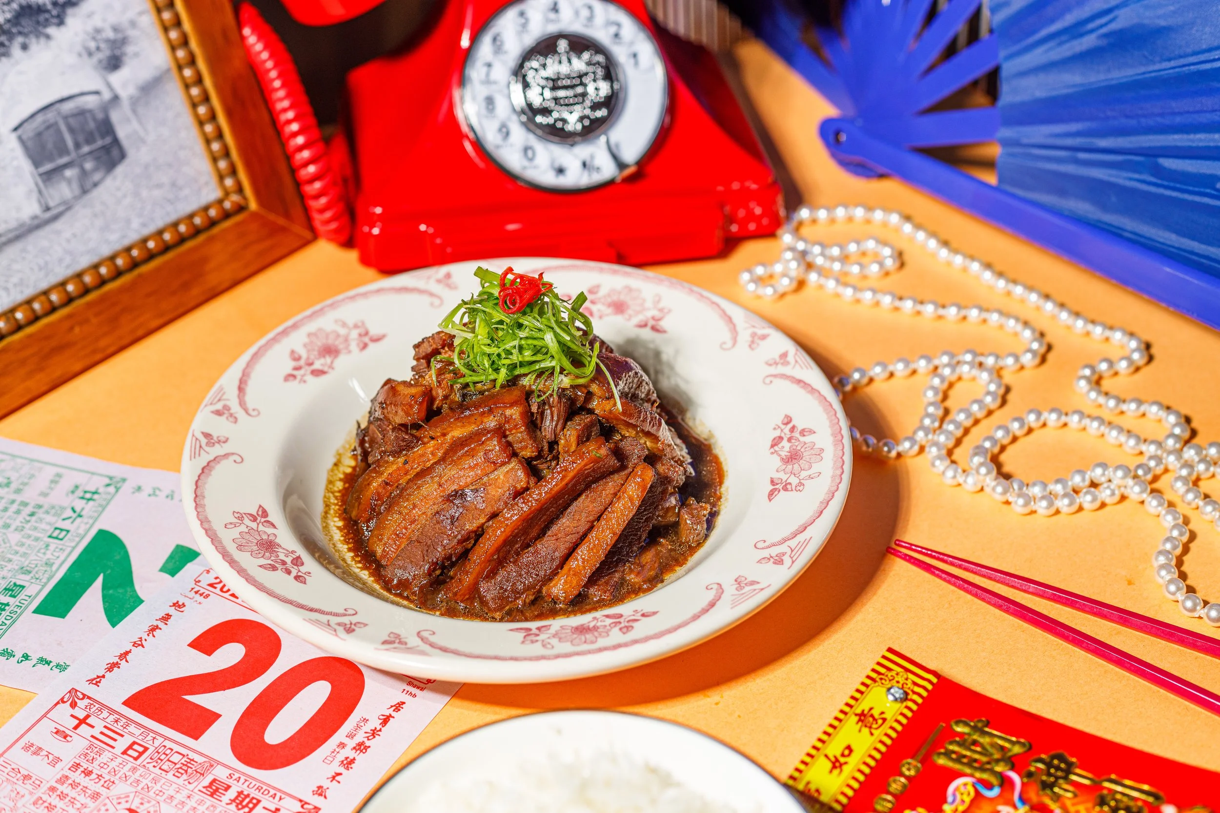 A plate of braised meat garnished with green herbs and red chili slices, placed on a table surrounded by Chinese cultural items, including a red vintage rotary phone, string pearls, red and yellow packets, and Chinese calendars.