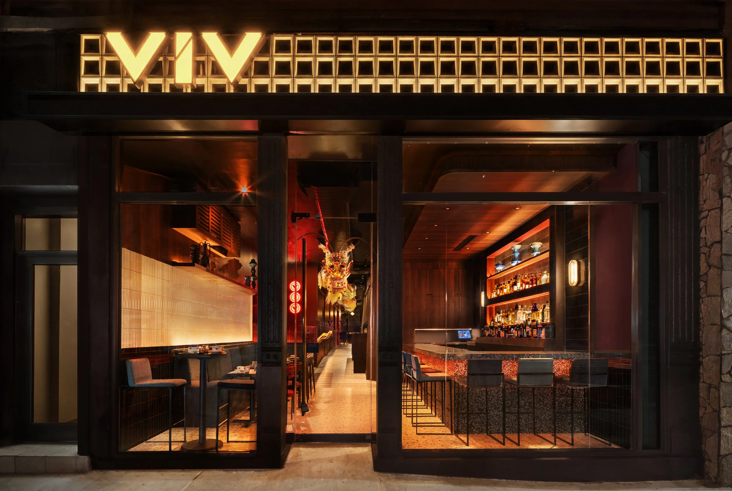 Night view of a modern bar or restaurant with illuminated signage reading 'VIVI' at the entrance, featuring a black and orange color scheme, bar stools, and interior lighting.