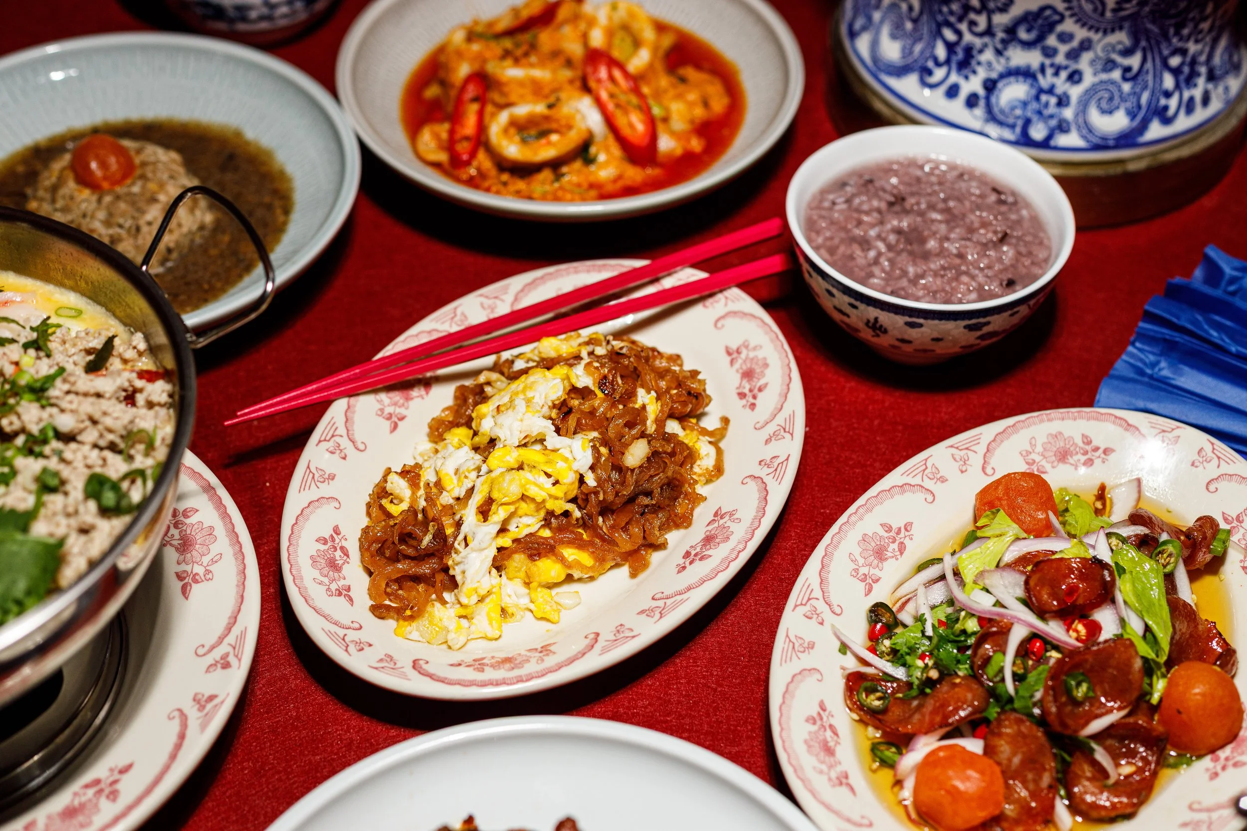Assorted dishes of Asian cuisine, including fried rice, noodle stir-fry, vegetable salad, and various hot and savory dishes on decorative plates and bowls, placed on a red tablecloth.