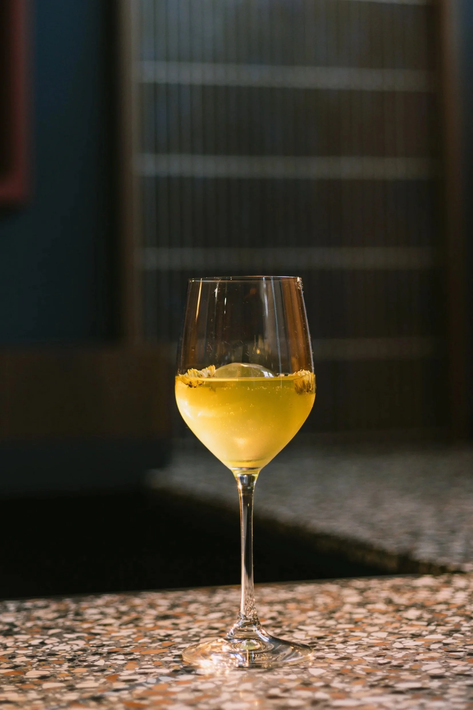 A glass of white wine with lemon and herbs inside, positioned on a patterned surface with a dark, blurred background.