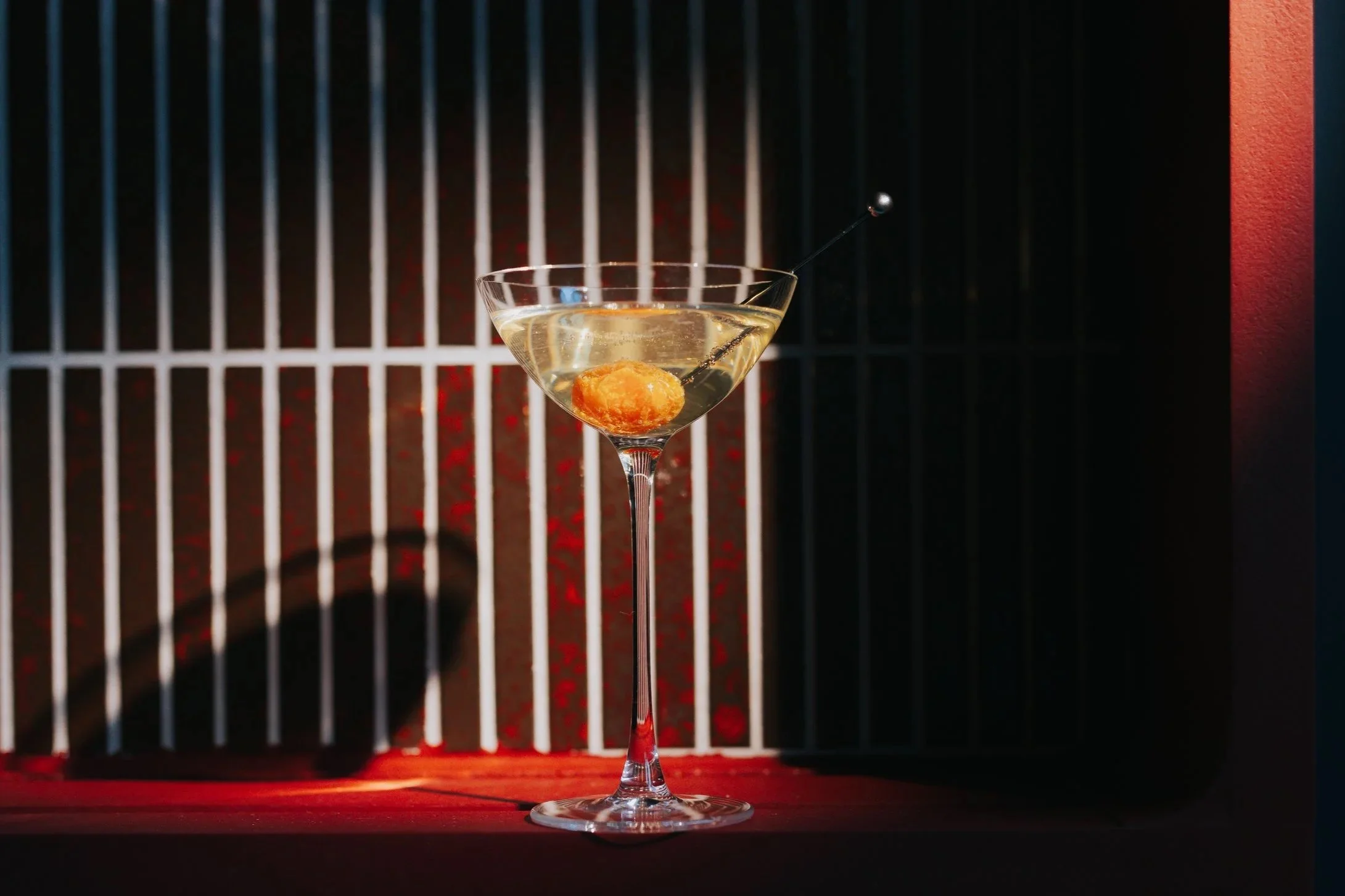 A martini glass filled with a clear beverage and garnished with a large golden cherry, with a black cocktail pick through the cherry, set against a background of vertical bars and dim lighting.