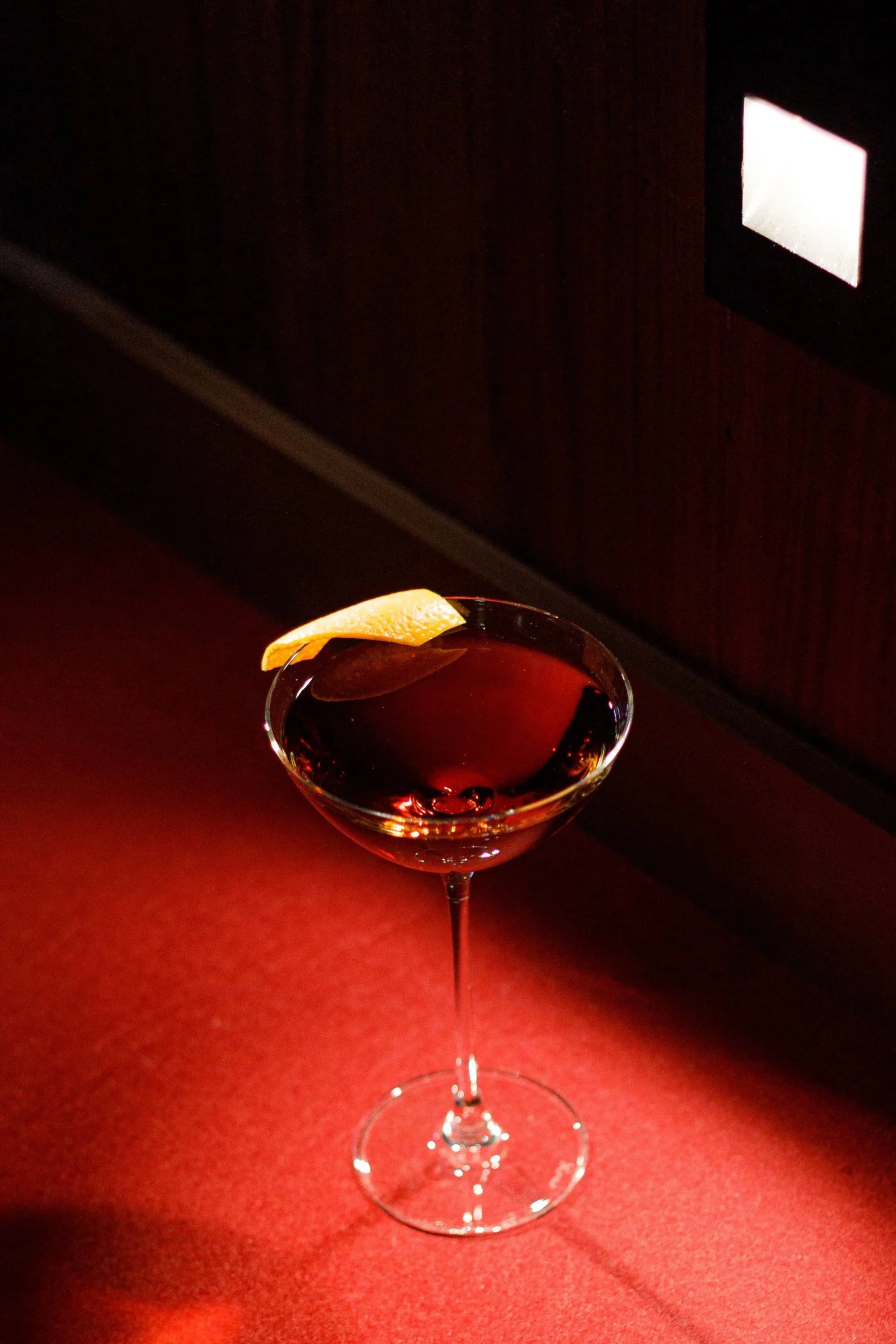 A cocktail glass with a dark red drink and a lemon twist garnish, set against a red background with a dark wall and wall sconce in the distance.