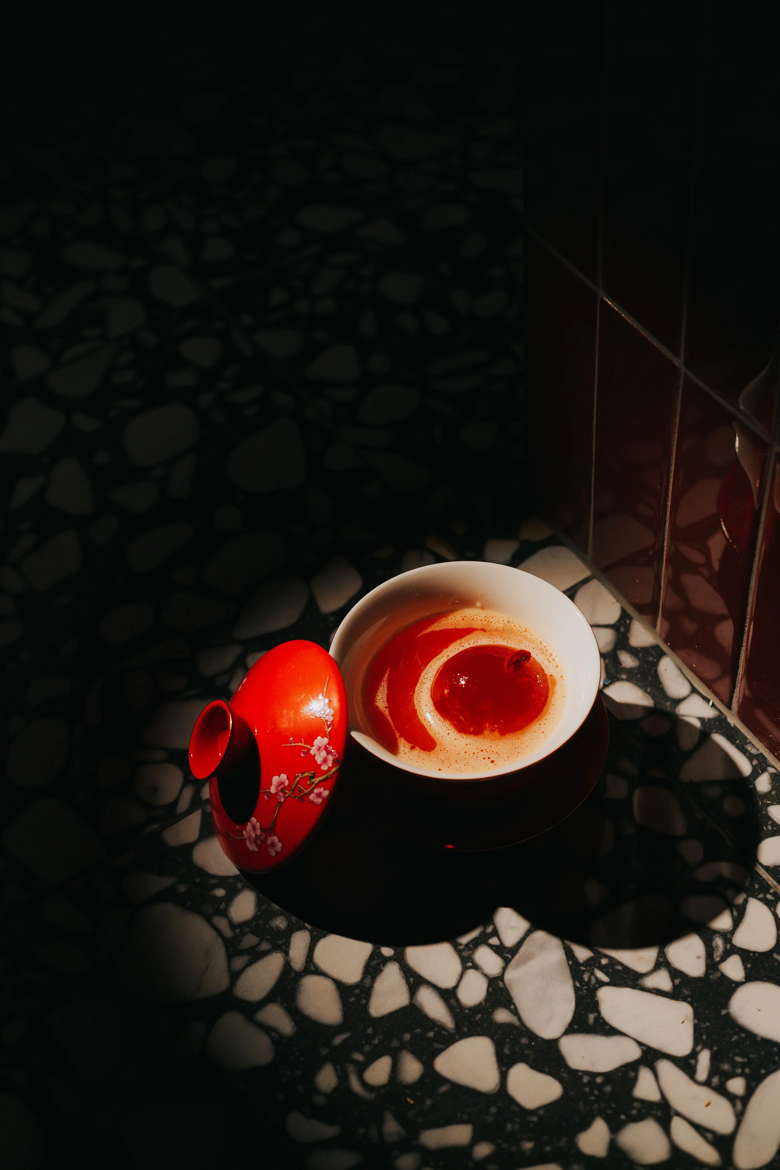 A white cup of coffee with a swirl of cream on top, placed on a dark plate with white irregular shapes. Next to the cup is a red decorative container with cherry blossom designs, and a red fruit, possibly a tomato, resting nearby. The background feat