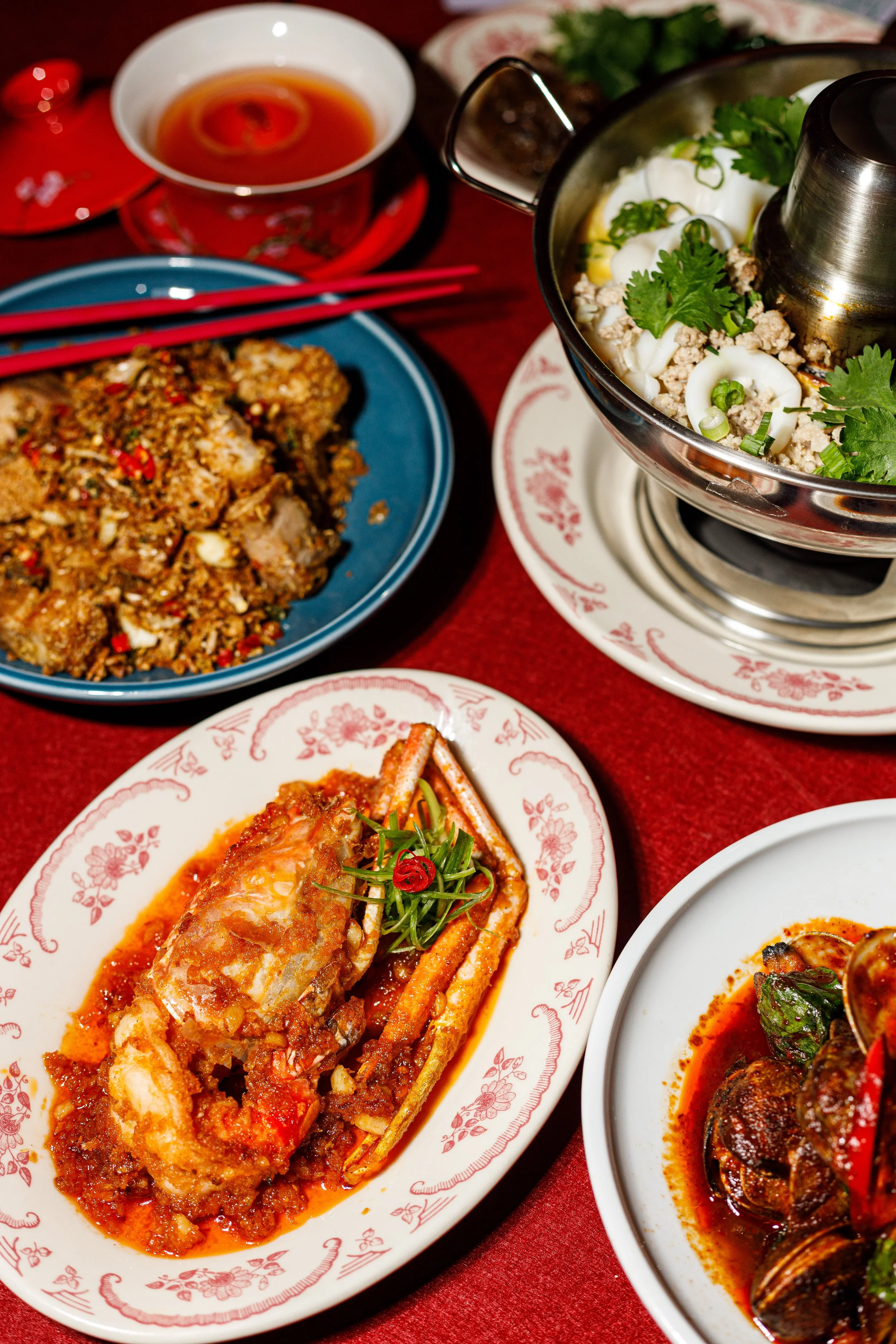 Close-up of Chinese dishes on decorative plates, including a steamed fish fillet with sauce, a meat and vegetable stir-fry, and a bowl of soup with herbs and cilantro, all on a red tablecloth with chopsticks and a dipping sauce in a bowl.