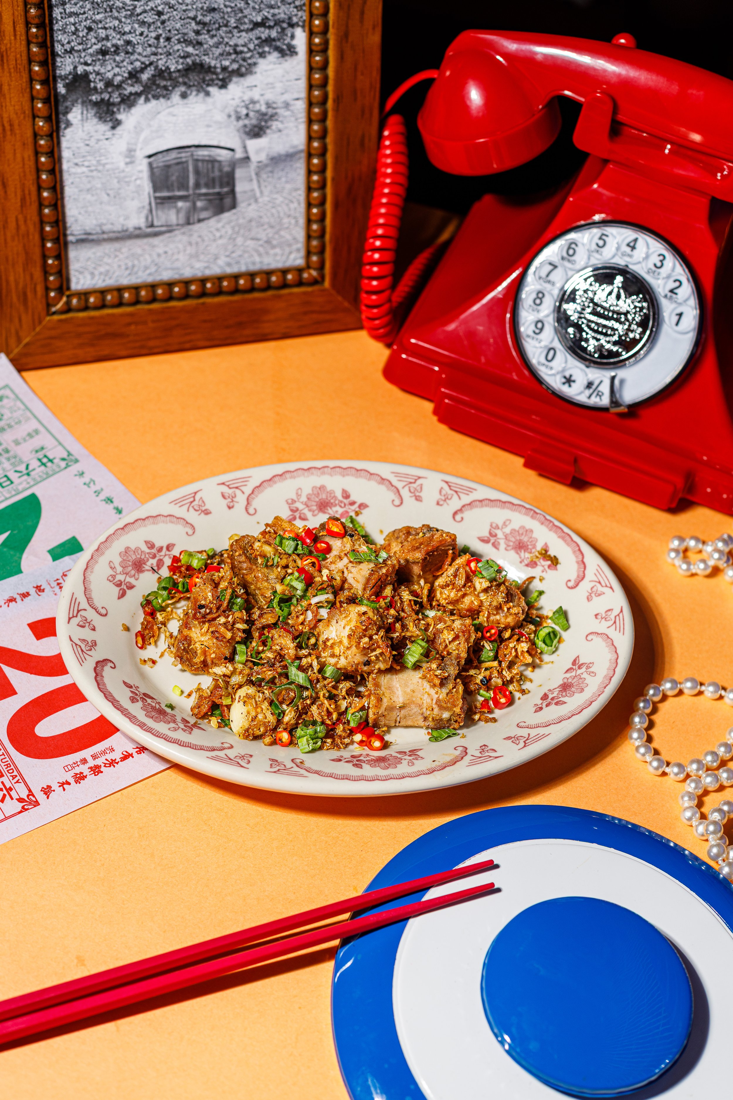 A plate of Chinese food garnished with chopped green onions on a table with a red vintage rotary phone, a framed black-and-white photo, red chopsticks, and a blue and white paper plate.