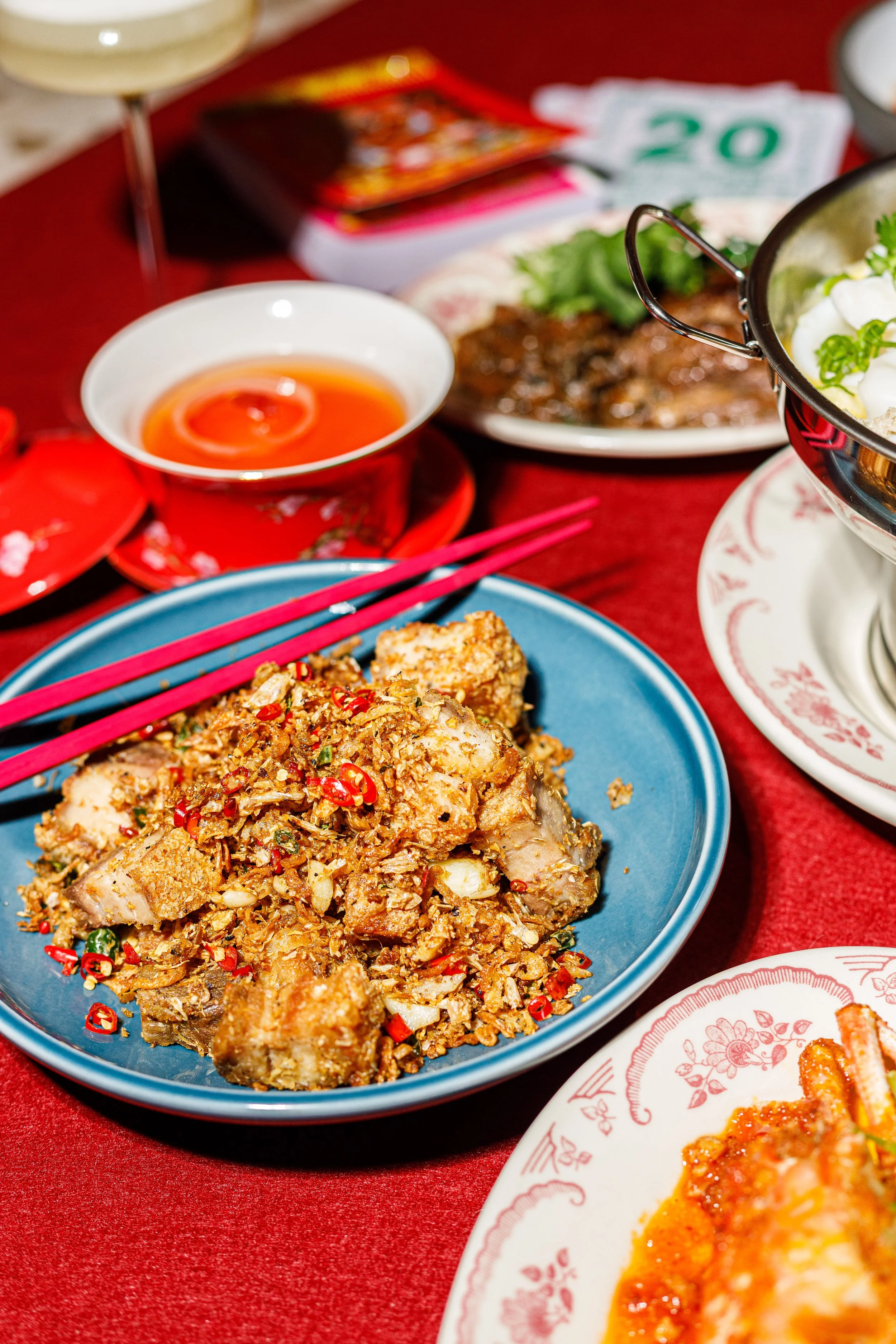 Close-up of a Chinese-style dish with chopped meat and vegetables, garnished with sliced red chili peppers on a blue plate. In the background, there are bowls with sauce and dishes with other food items, on a red tablecloth.