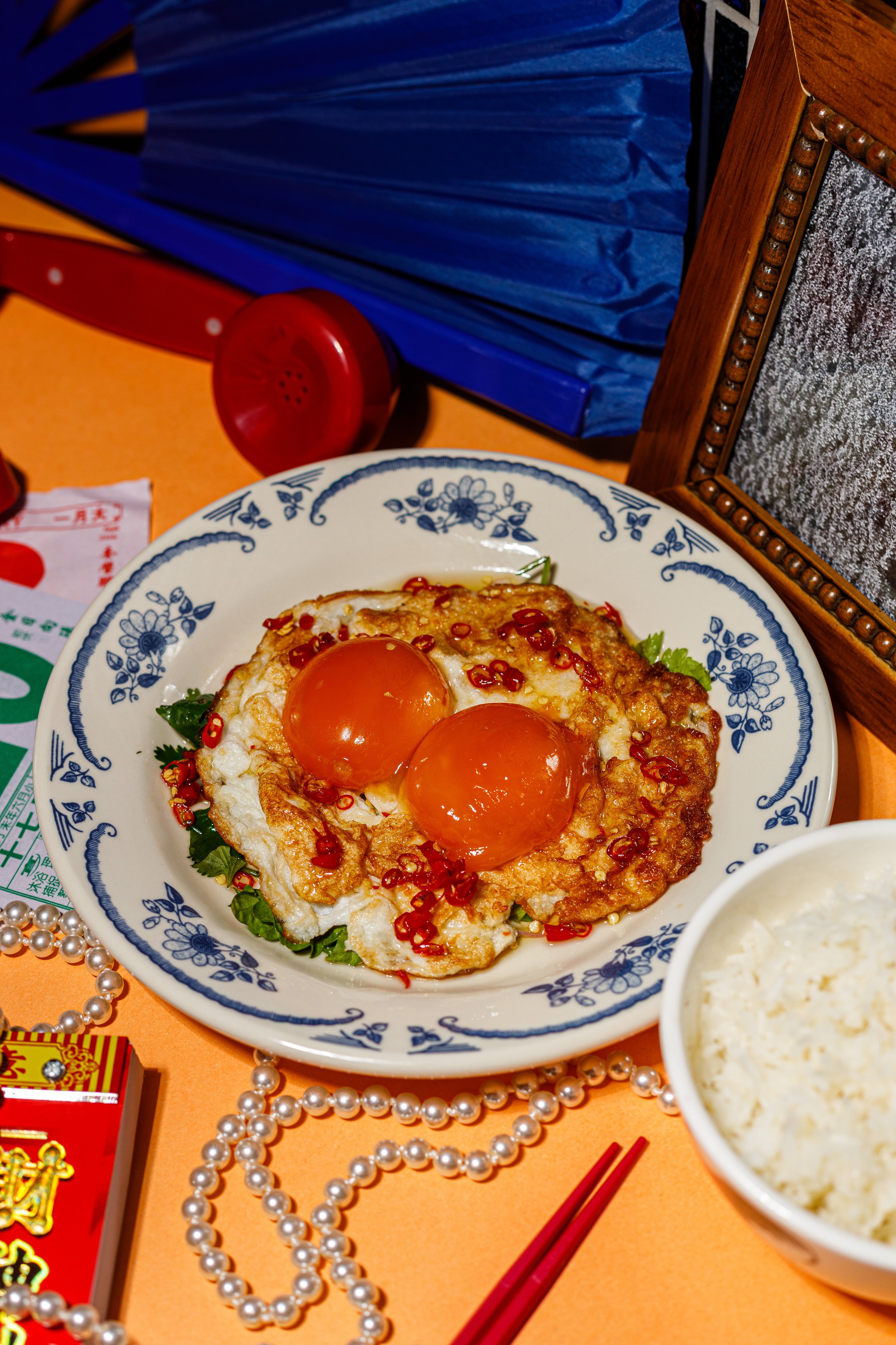 A plate with two sunny-side-up eggs garnished with red chili and surrounded by greens. The background includes a red telephone, pearl necklaces, red chopsticks, a bowl of rice, and festive decorations on an orange surface.
