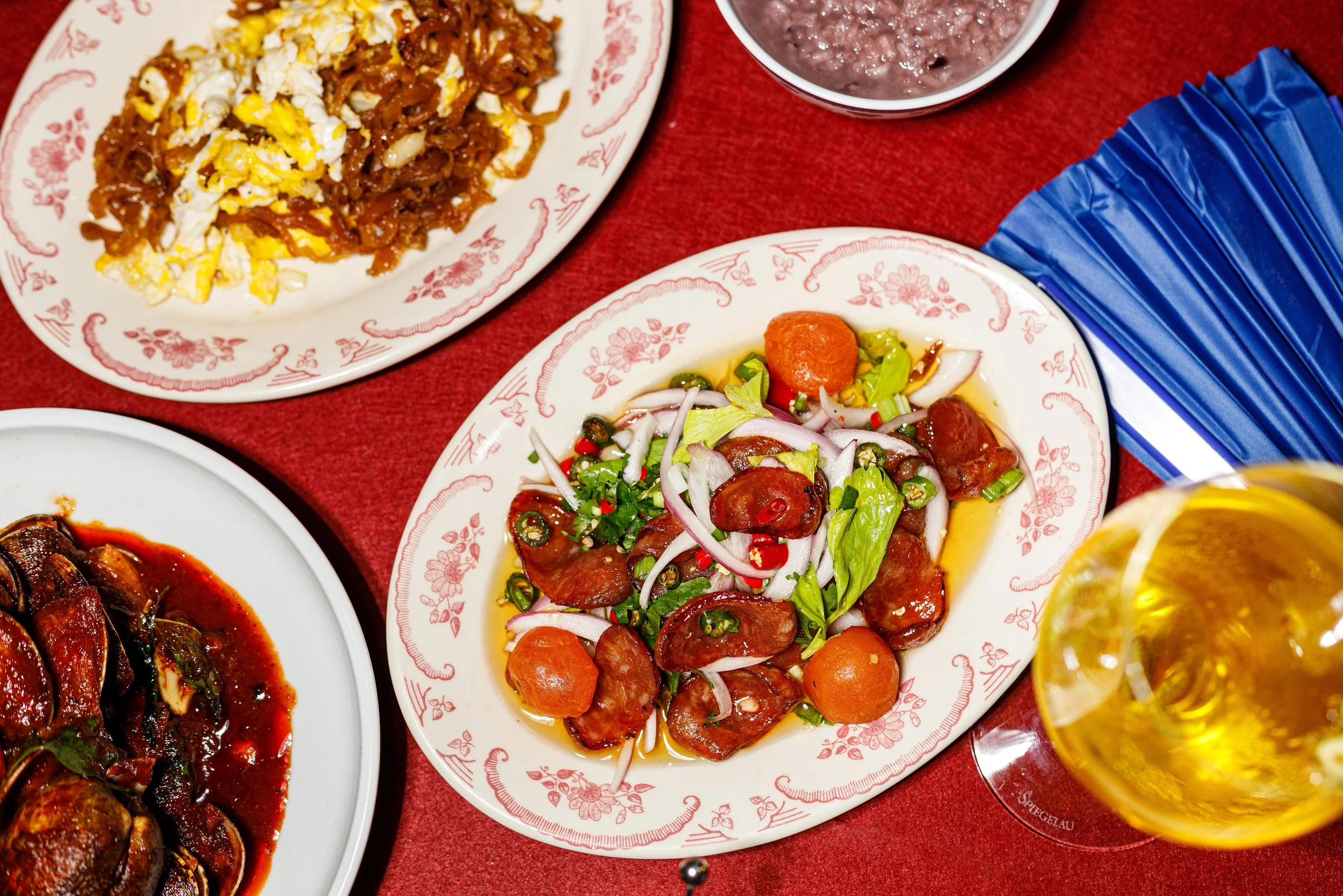 A table with Asian dishes on decorative plates, including a salad with sliced sausage, vegetables, and sauce, a plate of stir-fried noodles with scrambled eggs, a small bowl of purple rice pudding, and a glass of beer.