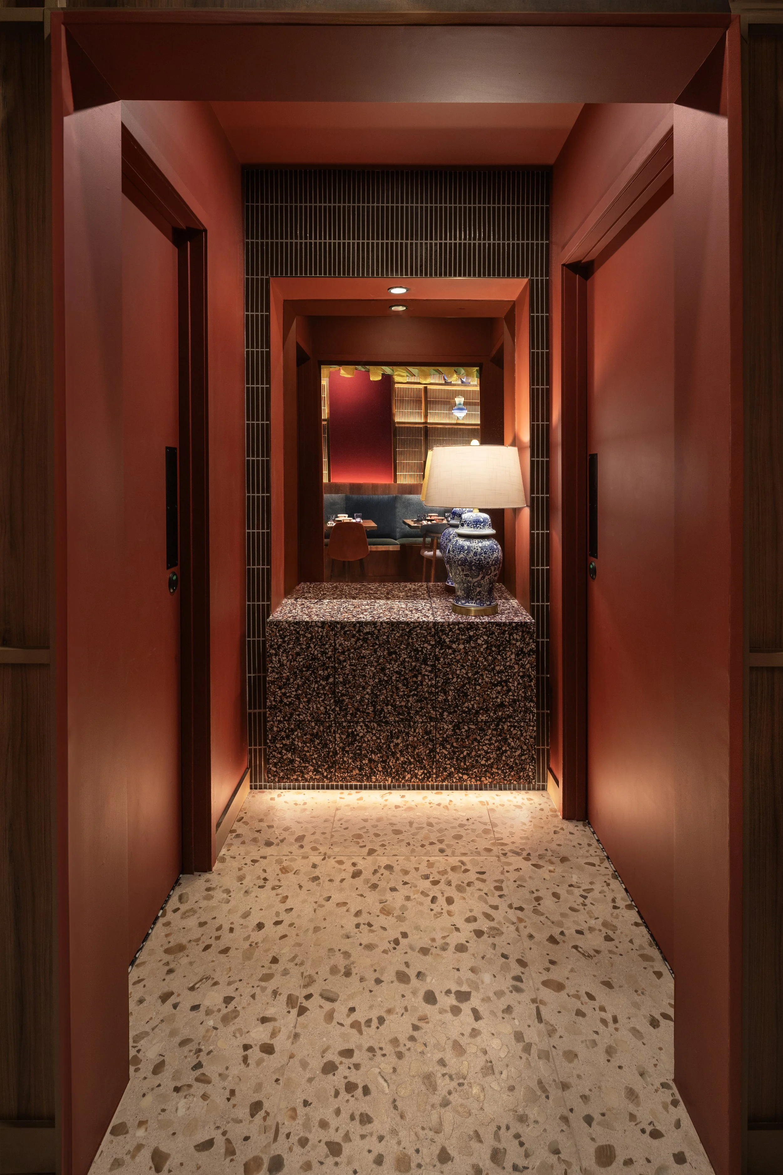 View through a red-walled corridor leading to a furnished room with a lamp on a patterned table, chairs, and shelves with decorative objects.