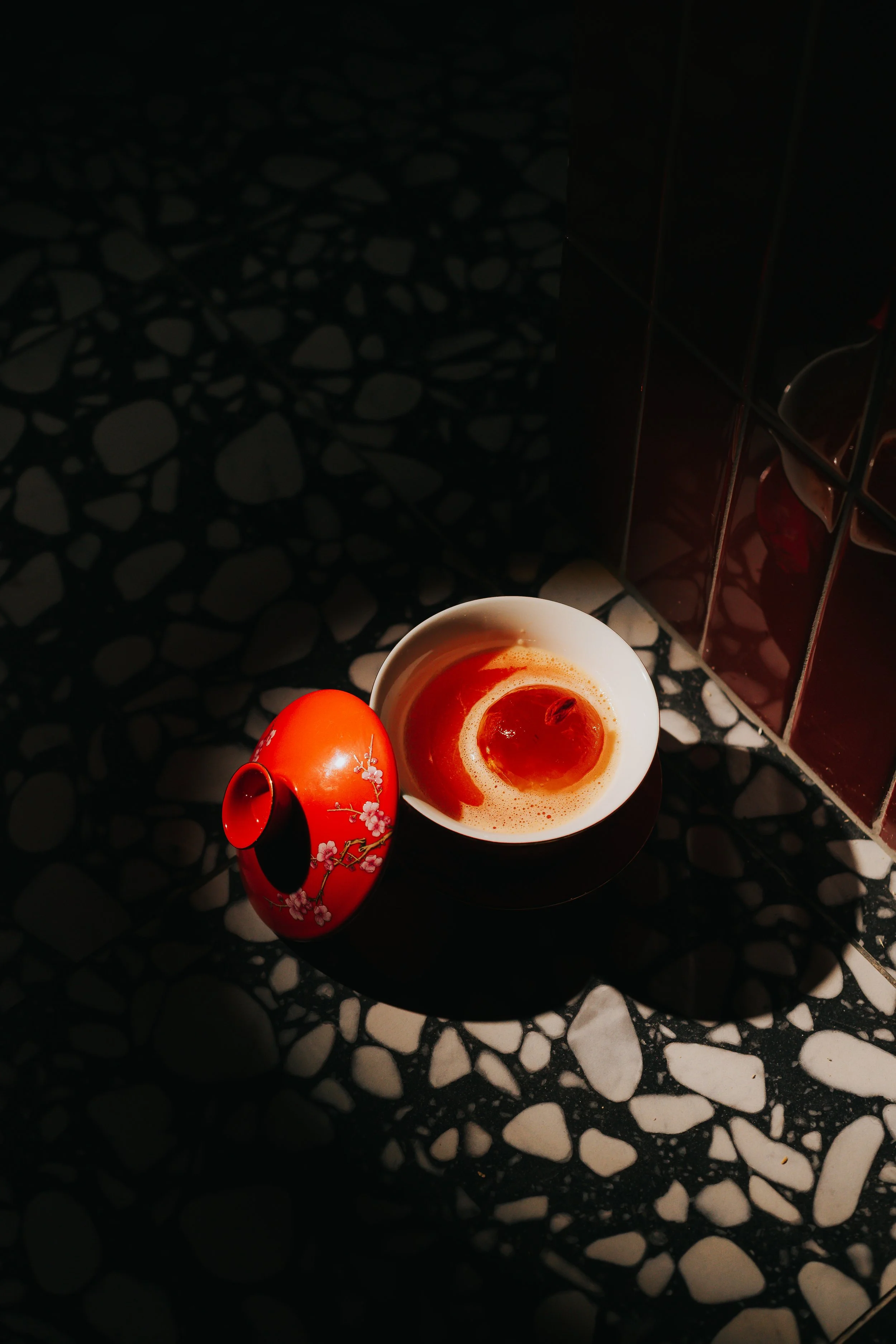 A decorative red container with floral patterns and a black lid next to a white bowl with a dessert topped with a cherry, placed on a black and white pebble-patterned surface, with a dark tiled wall in the background.