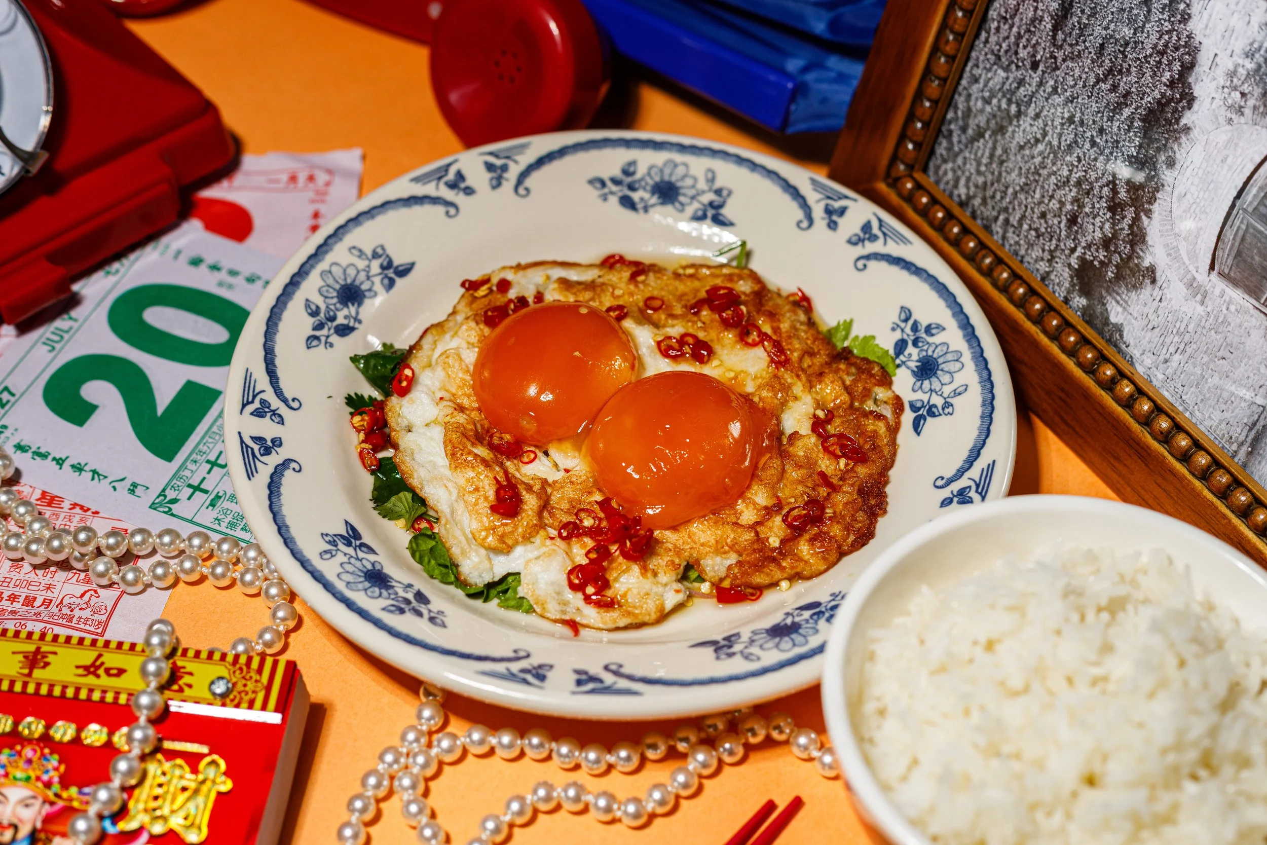 A plate with two sunny side up eggs garnished with red pepper flakes and some green herbs, placed on a table surrounded by various objects including red and white lottery tickets, pearl necklaces, a red toy phone, a framed autumn landscape photo, a bowl of rice, and some colorful packaging.