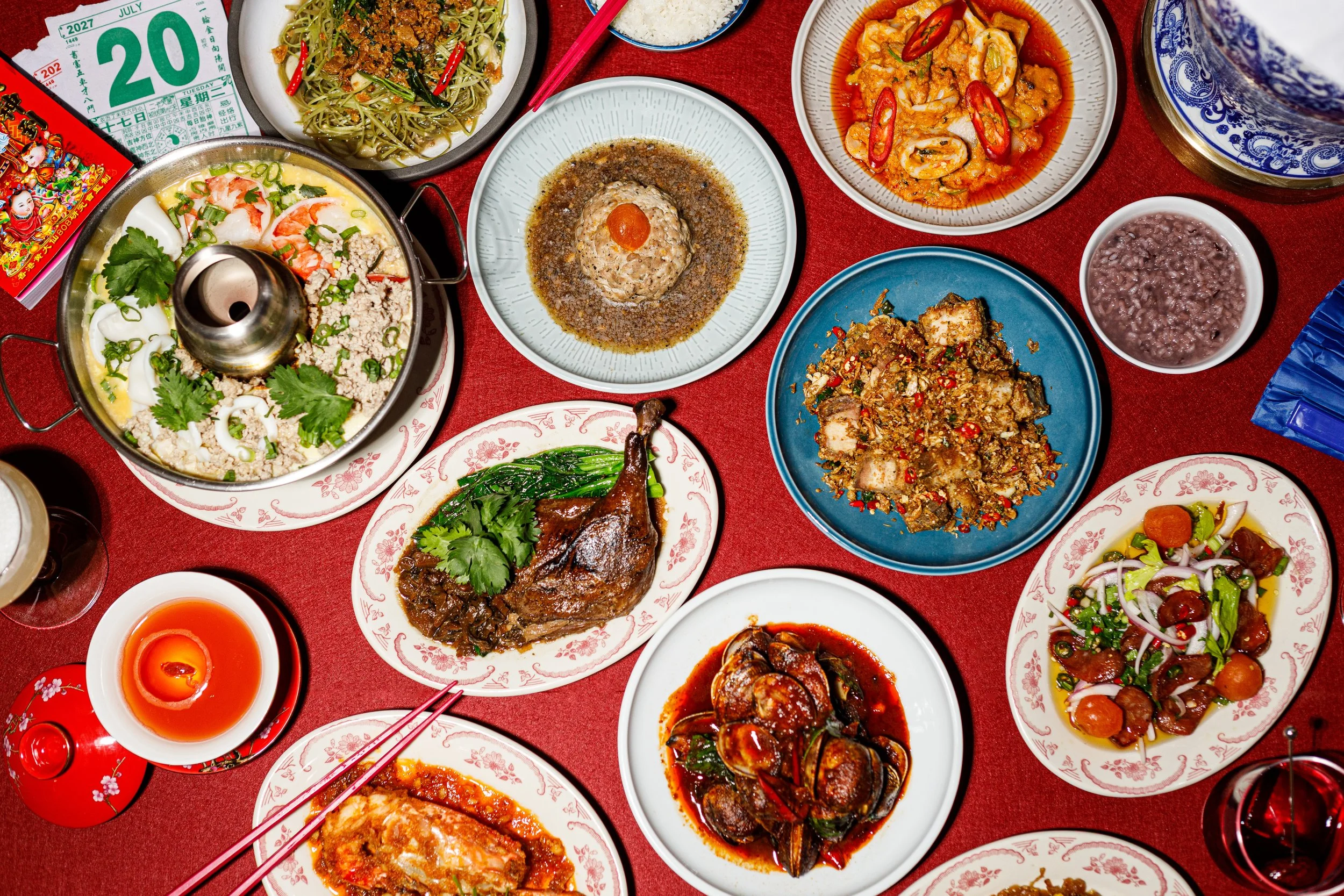 A variety of Chinese dishes served in bowls and plates on a red tablecloth, including stir-fried vegetables, fish, meats in sauces, rice, and a tea set.