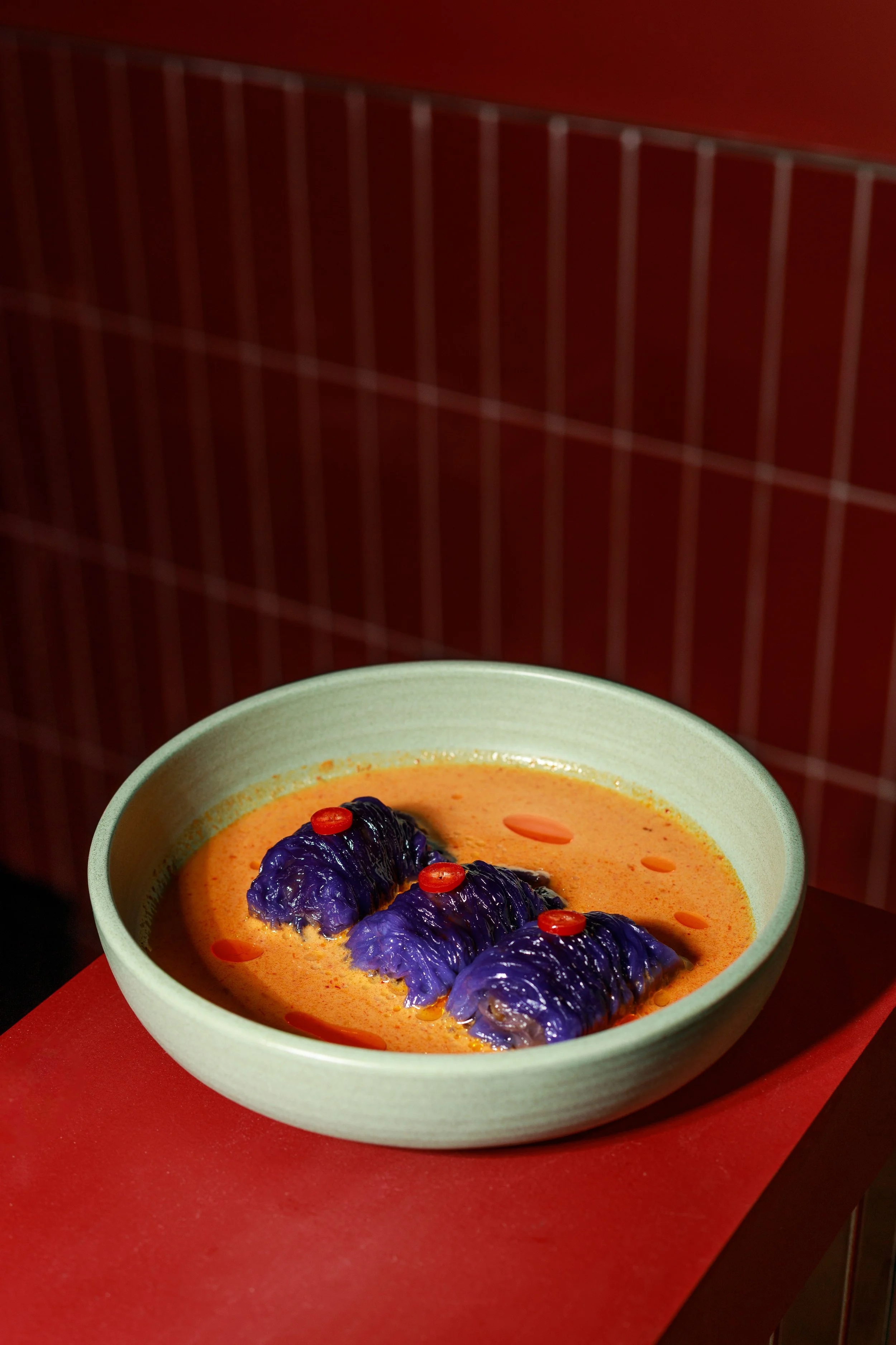 A bowl of orange-colored soup with purple eggplant and red pepper slices as garnish, placed on a red surface with a dark background.