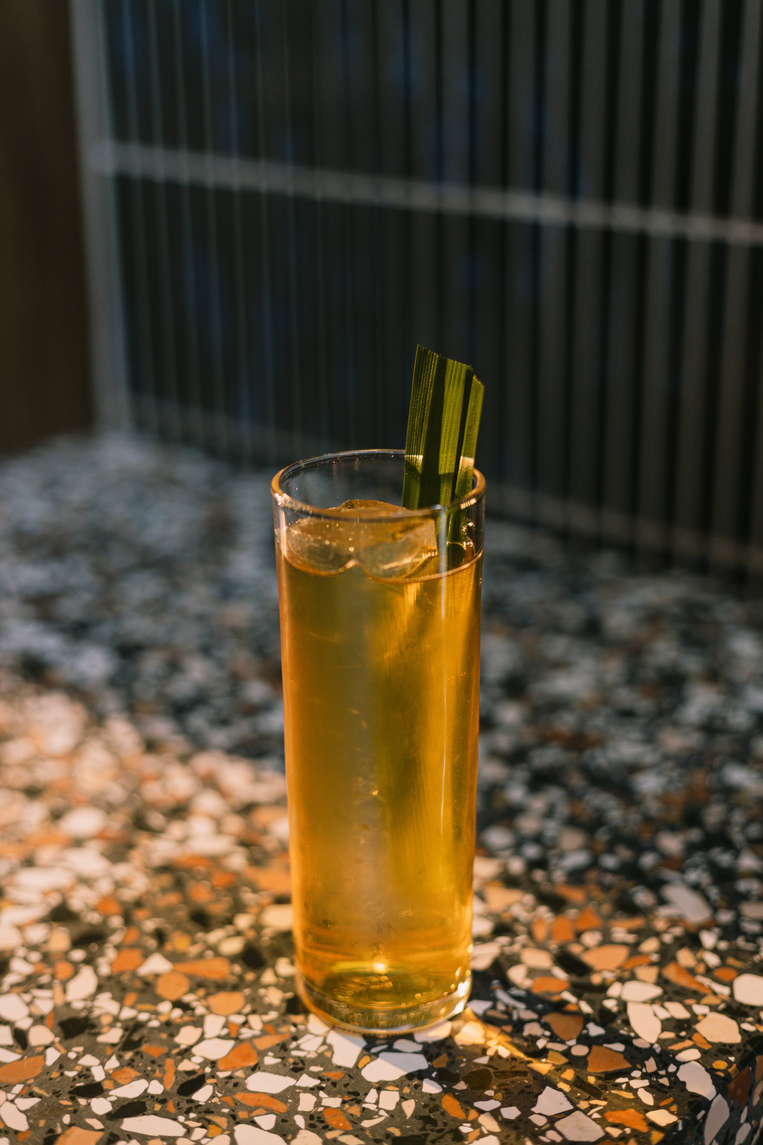A tall glass filled with a yellowish drink and ice, garnished with two green strips of lemongrass in a dimly lit setting.