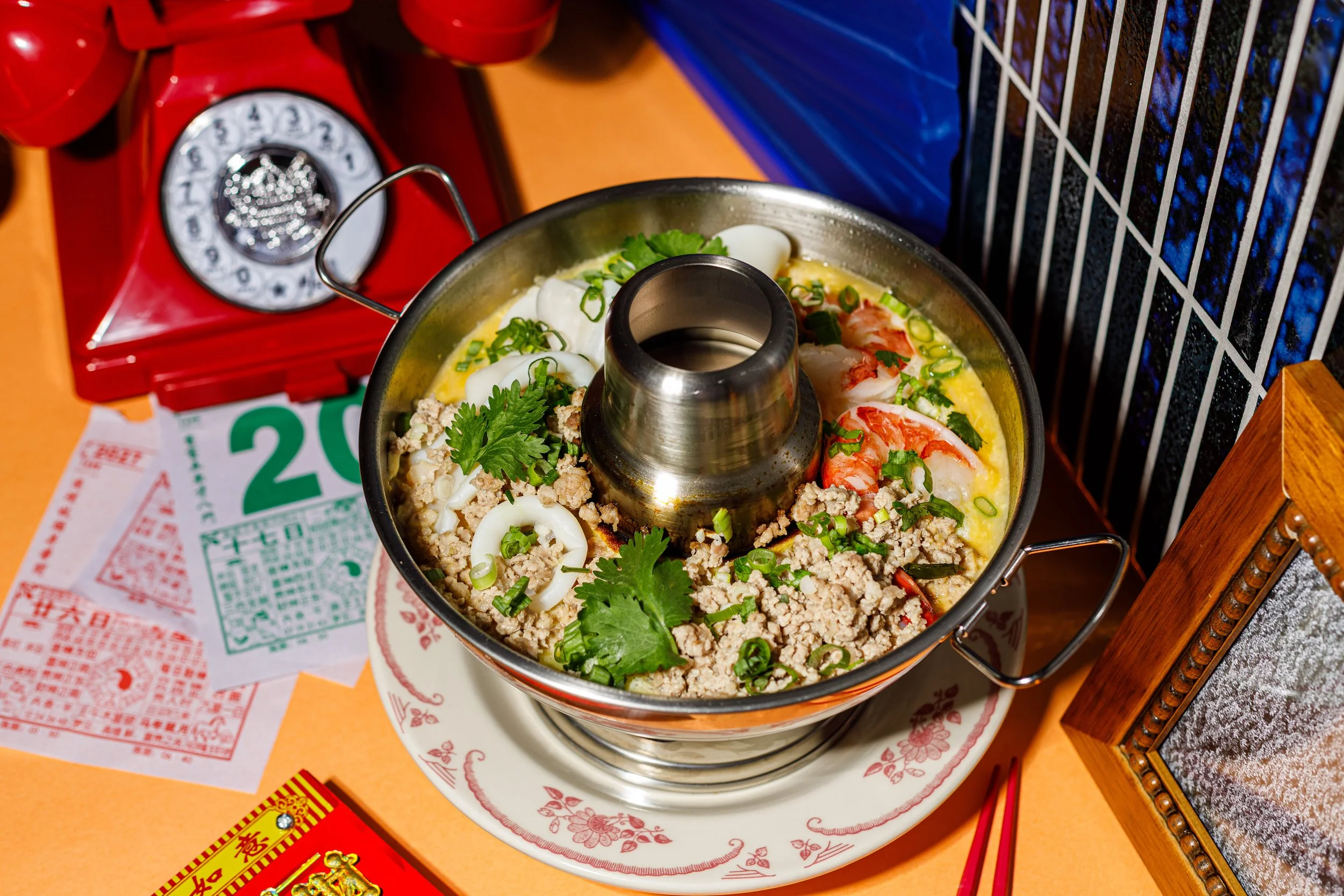 A hot pot filled with chopped green onions, cilantro, minced meat, shrimp, squid, and boiled eggs on a table with a red rotary phone, lottery tickets, and a red box of Chinese snacks nearby.