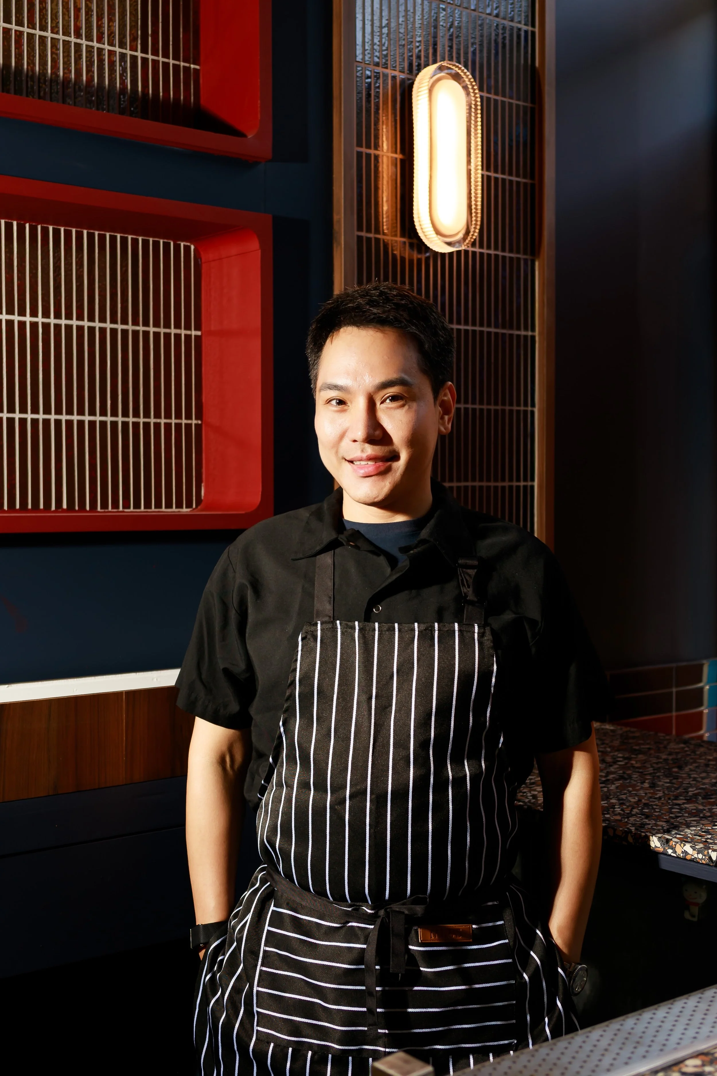A young man wearing a black shirt and a black-and-white striped apron stands in a restaurant or café, smiling at the camera. The background features decorative wooden and metal wall panels with a warm glow from a modern wall-mounted light fixture.