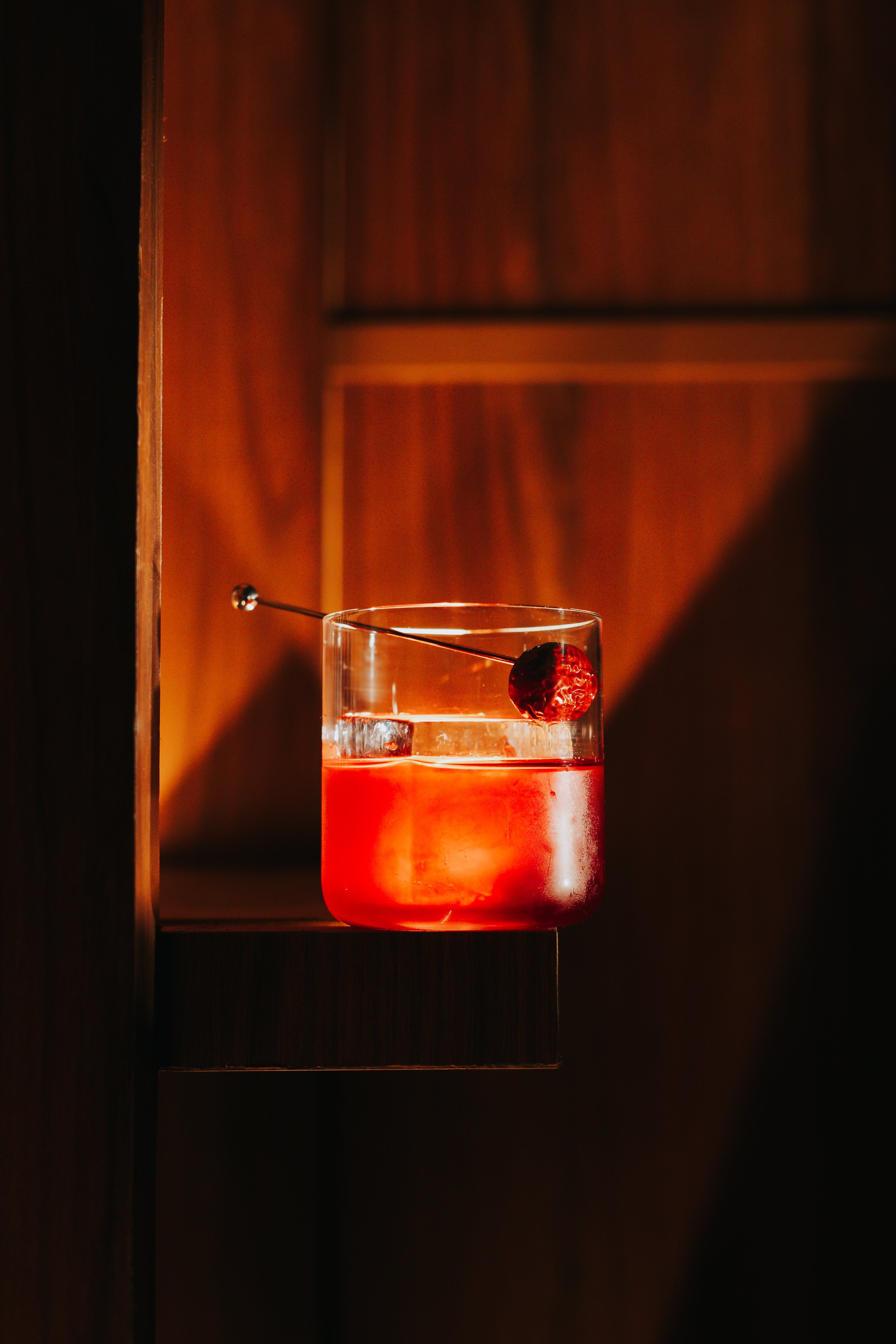 A glass of red cocktail garnished with a cherry and served with ice, placed on a dark surface with warm lighting.