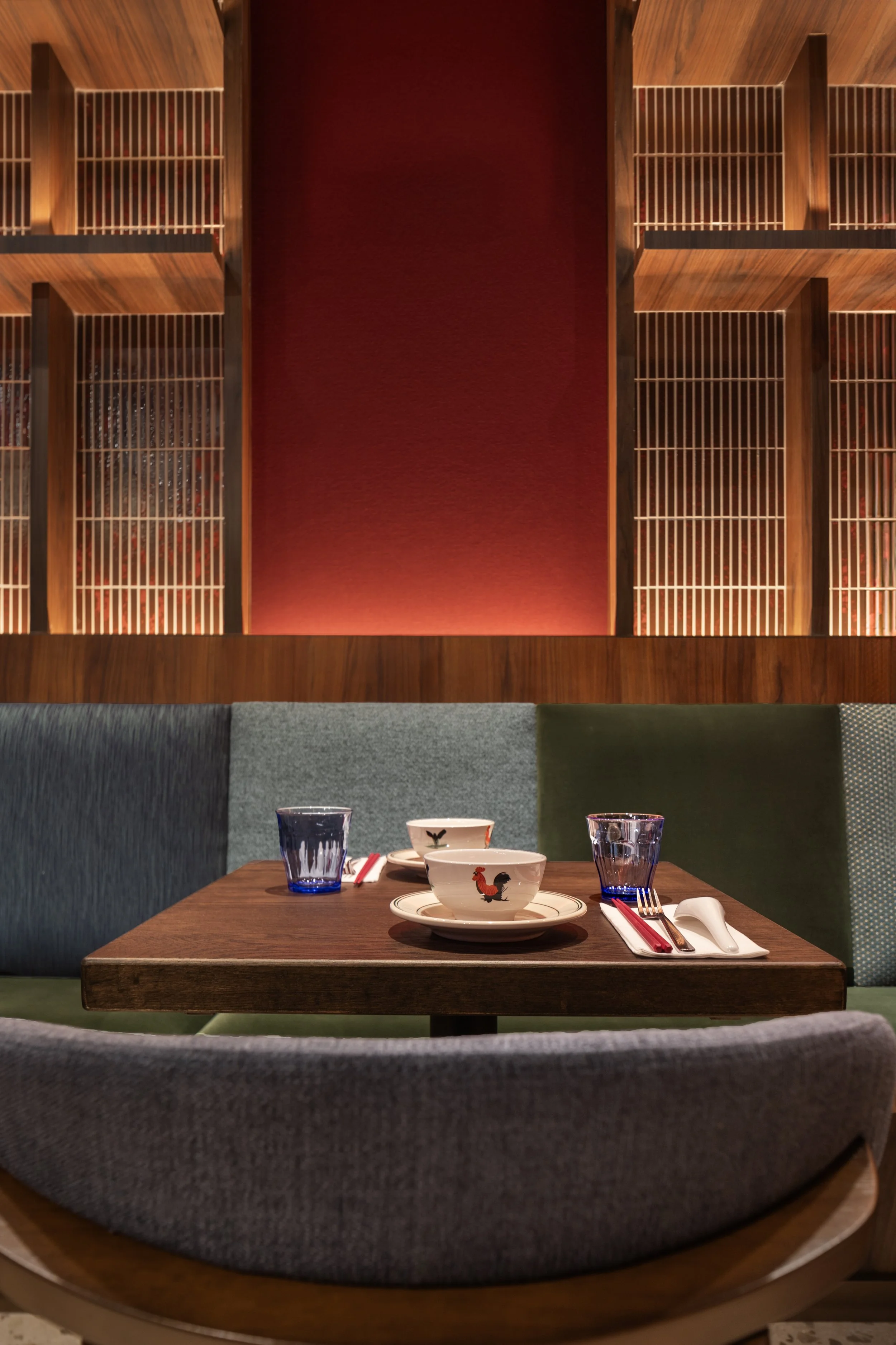 A table set for two in a restaurant with a matching porcelain bowl, chopsticks, glasses, and forks, with a colorful upholstered booth in the background and a decorative wooden wall with red cloth paneling above.