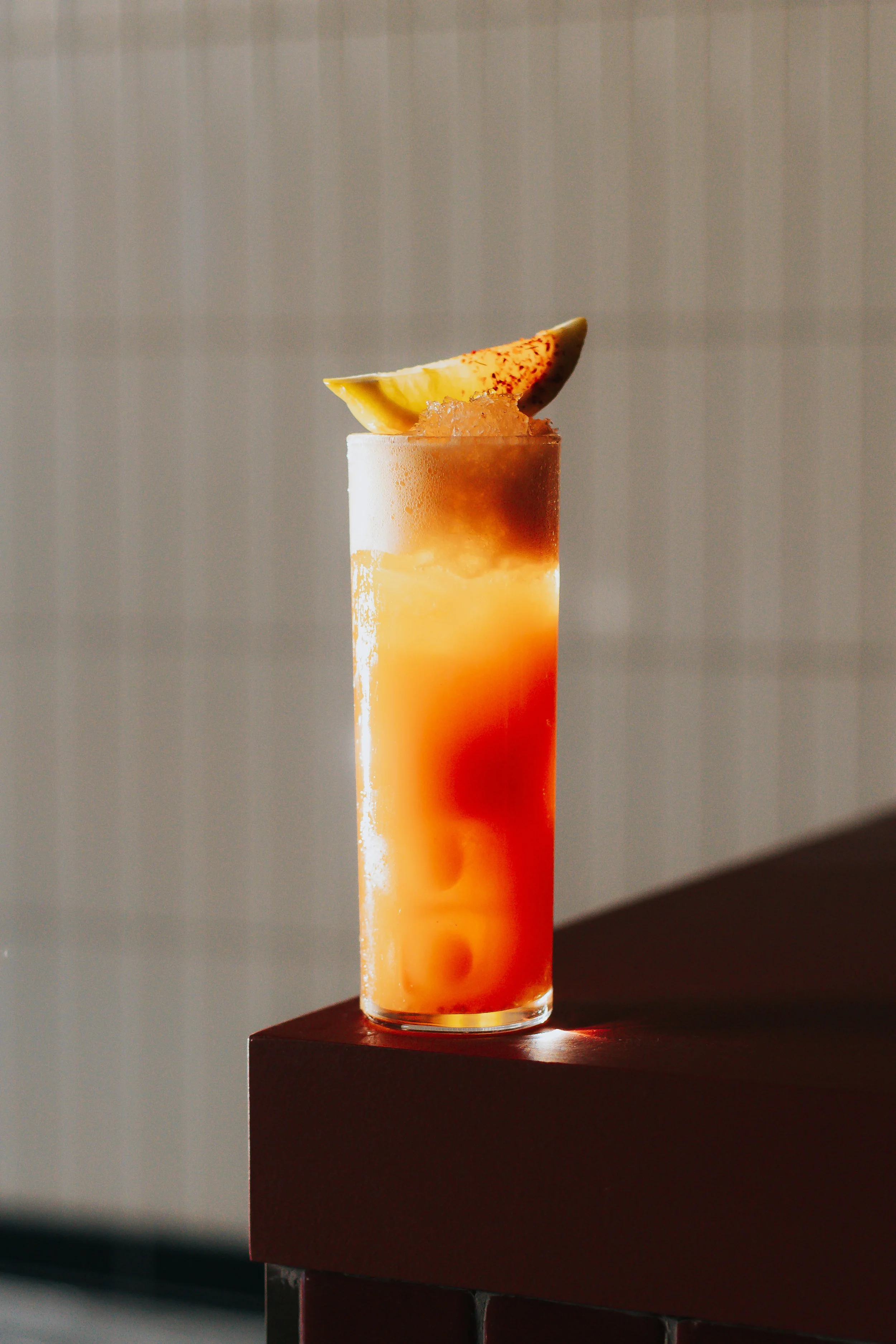 A tall glass of a layered orange and red beverage topped with a lemon wedge garnished with red spice powder, placed on a dark table.