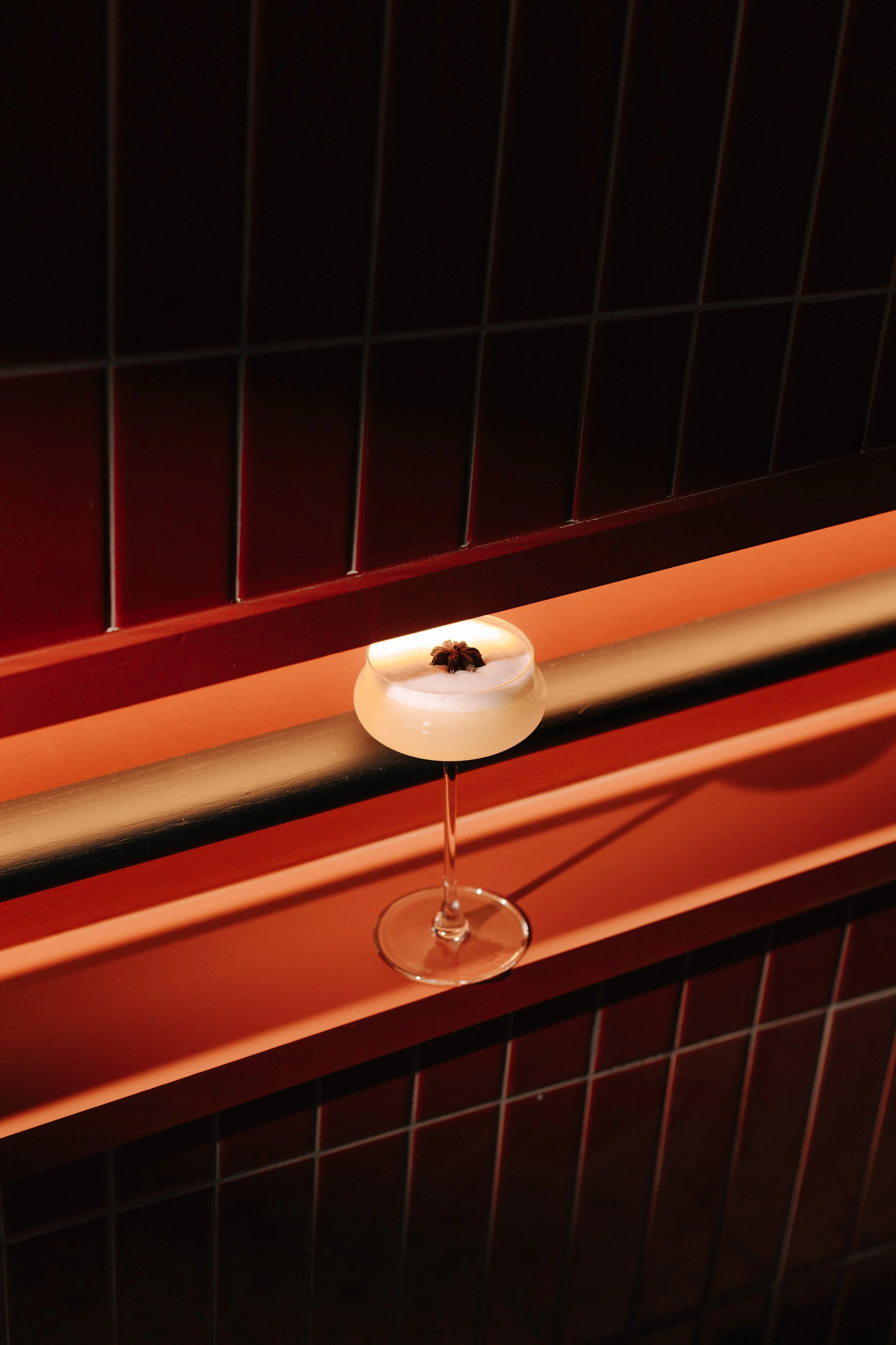 A cocktail glass with a frothy white drink topped with a small flower, placed on a wooden surface in front of a black tiled wall.