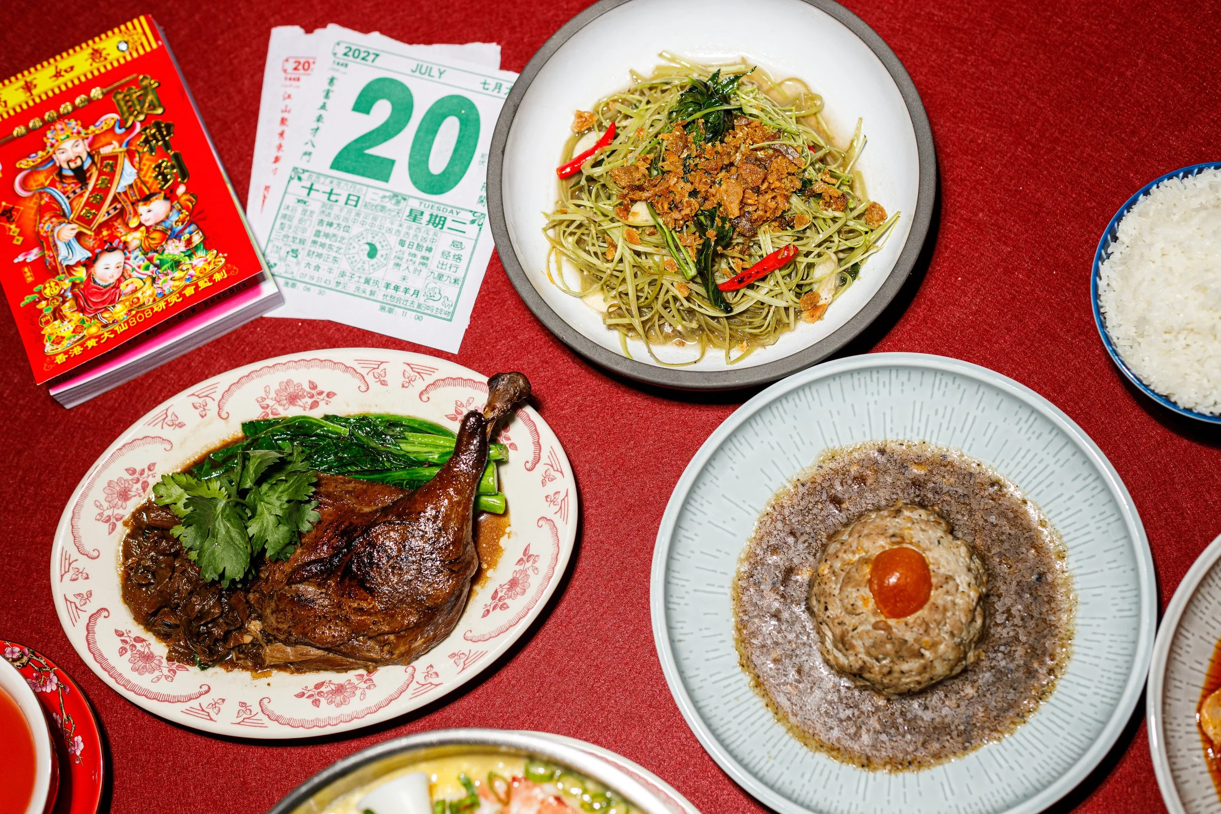 A table with various Asian dishes, including a plate of noodles with vegetables and chili peppers, a bowl of rice, a plate with a cooked chicken leg topped with herbs, and a dish with a spiral of seasoned meat or fish, alongside red envelopes and a c