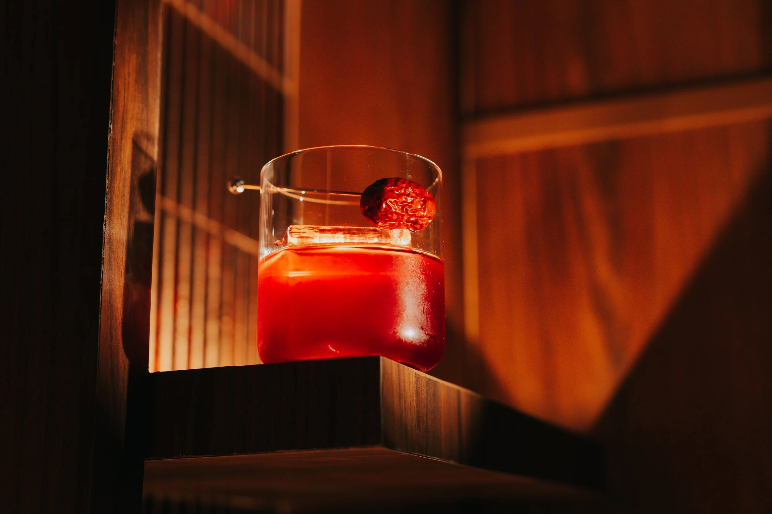A glass with a red beverage garnished with a red date fruit on the rim, placed on a wooden surface with warm lighting