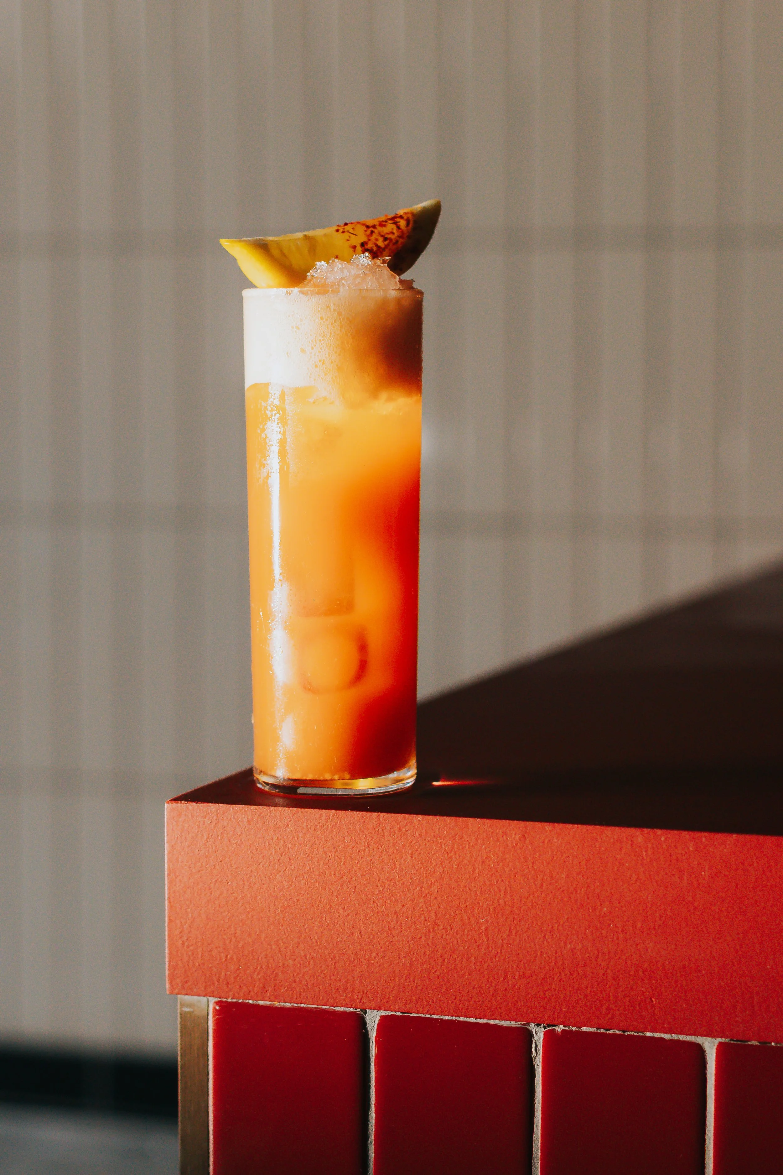 A colorful cocktail with a pineapple slice garnish, placed on an orange platform with a gray striped background.