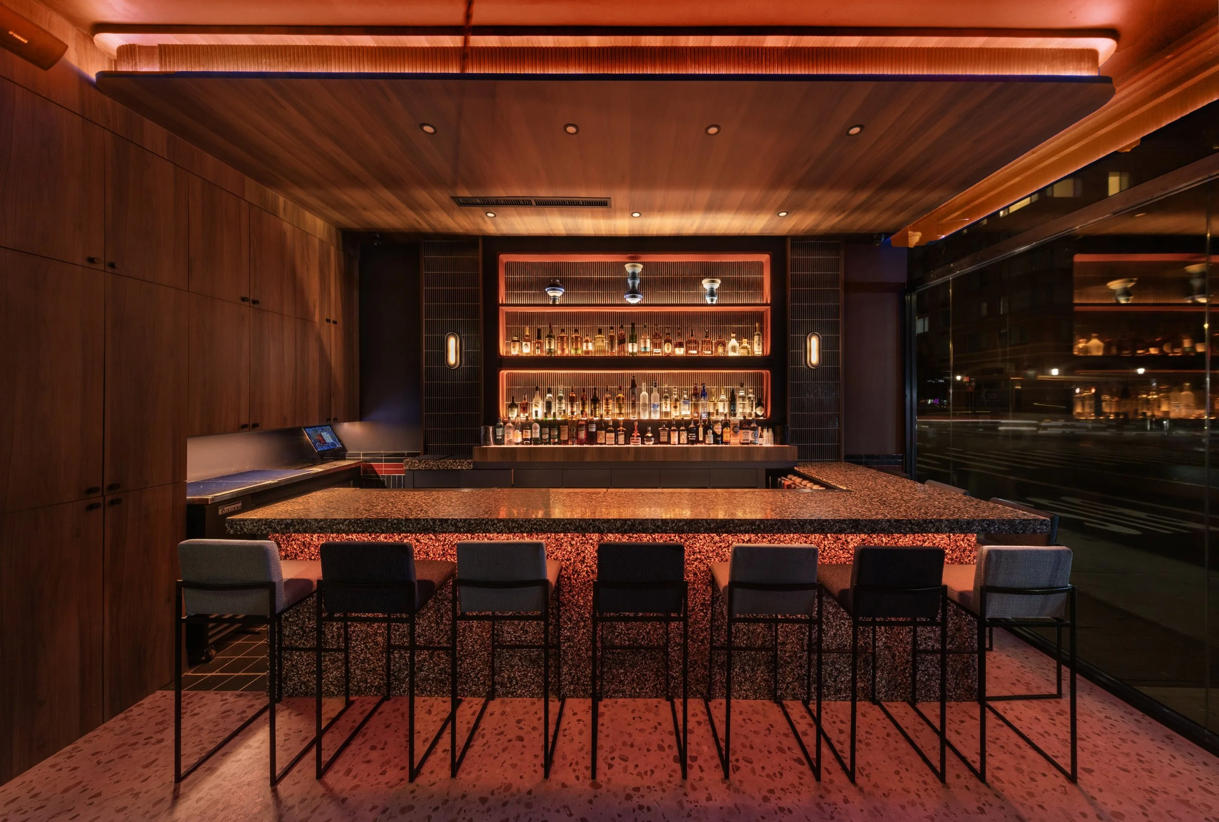 A bar area with a wooden ceiling and walls, illuminated shelves with bottles of alcohol, a granite counter, and chairs facing the bar.