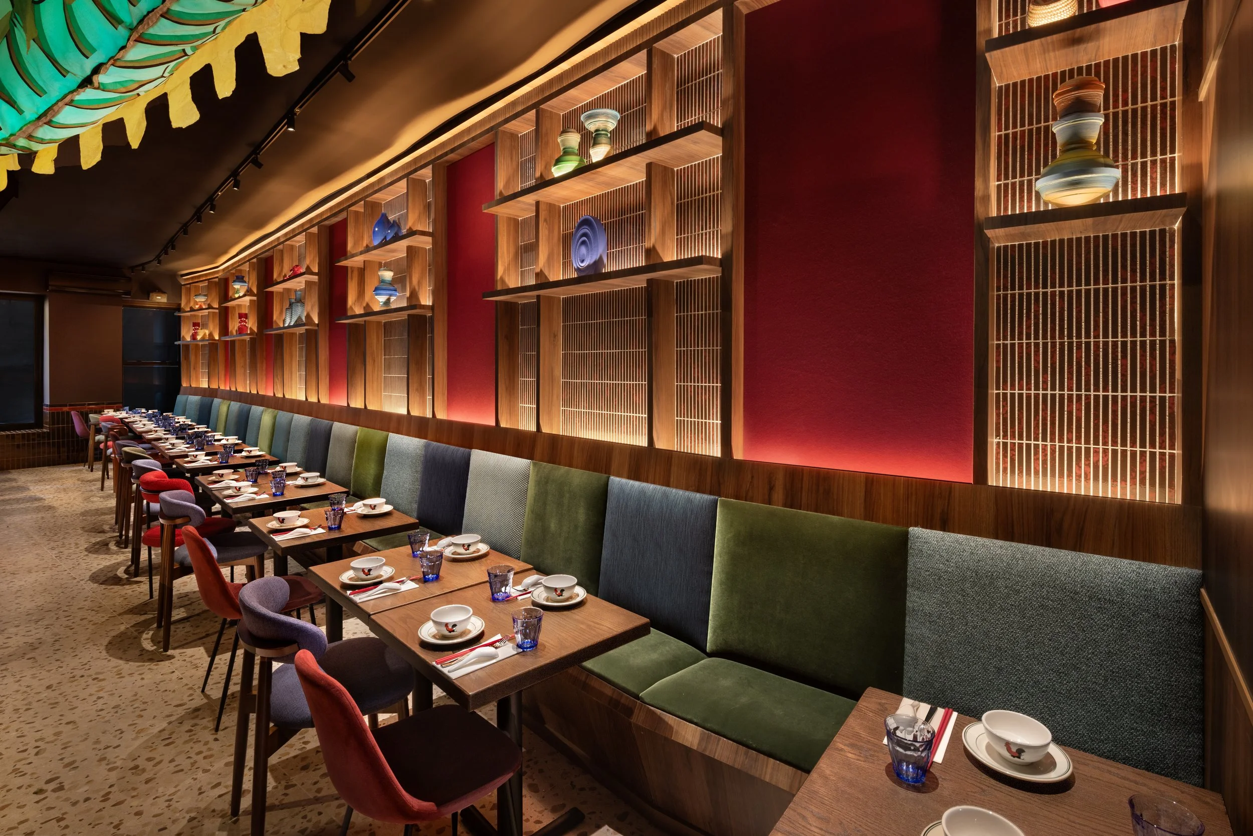 A restaurant dining area with a long wooden table set with bowls, glasses, and utensils. Colorful chairs surround the table. A decorative wooden wall with illuminated shelves holding colorful pottery and decorative objects is visible. The ceiling has