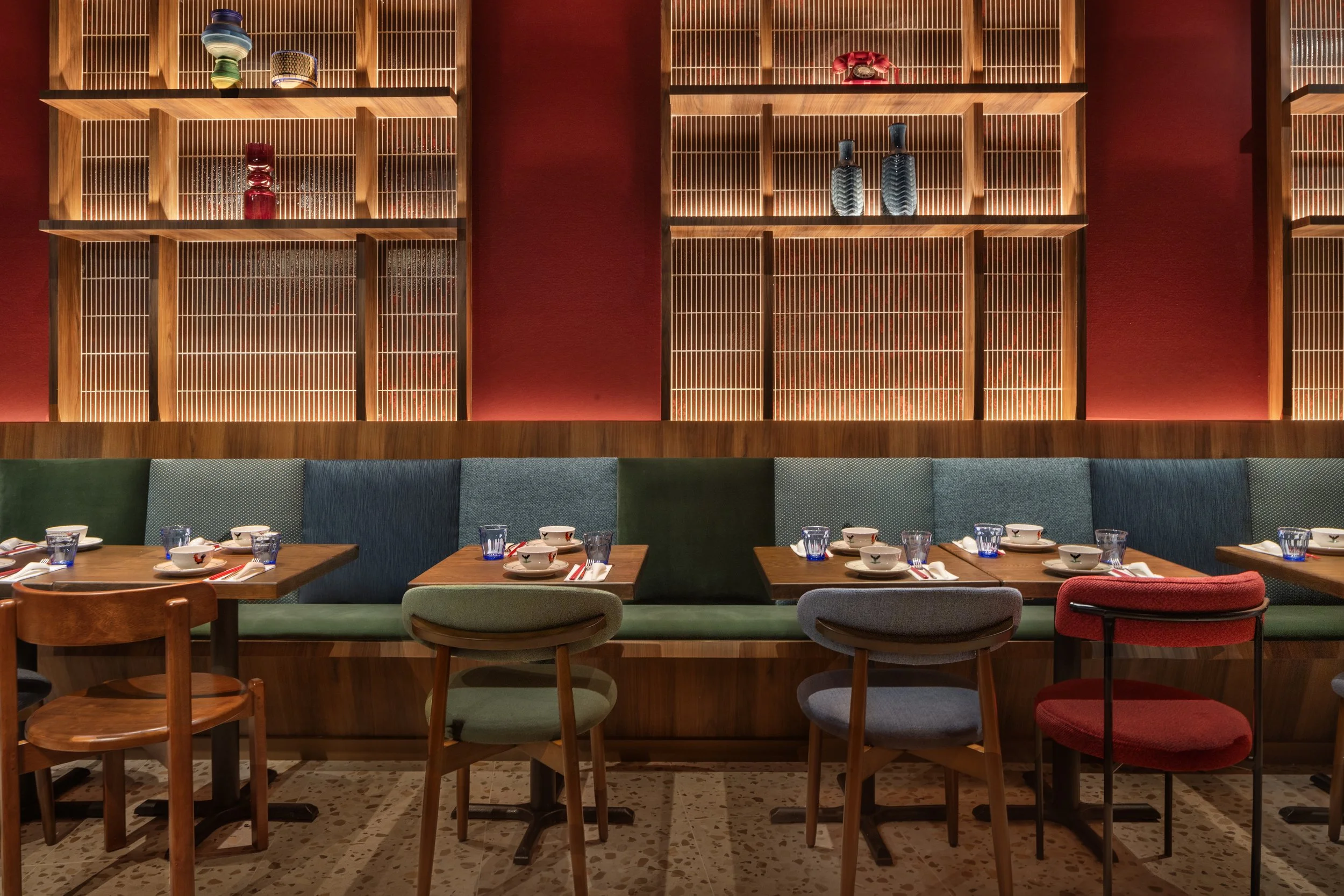 A restaurant interior with wooden slat wall panels and warm lighting. There is a row of tables set with bowls, glasses, and utensils, and a cushioned bench seating against the wall featuring different colored and patterned chairs.