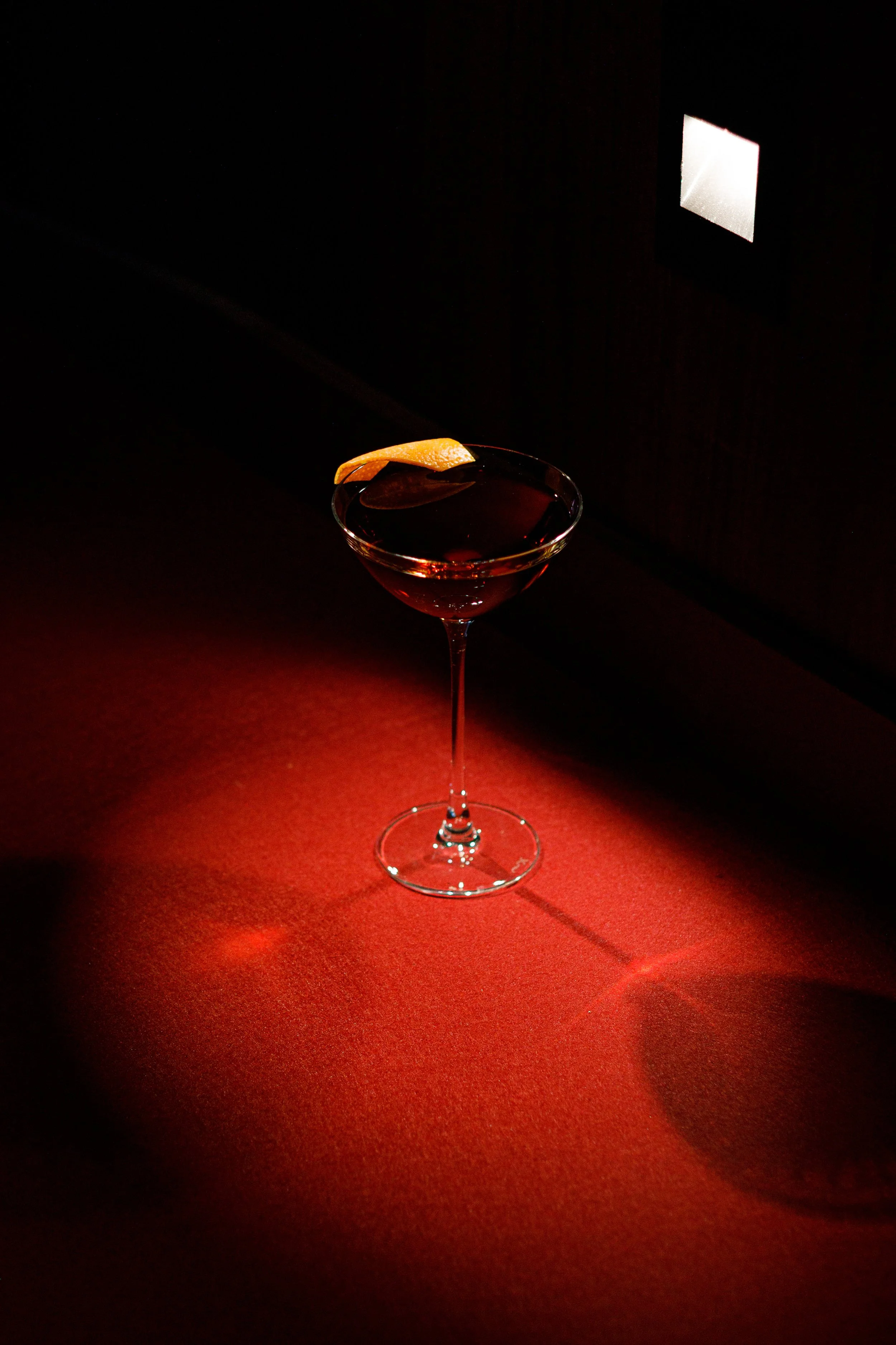 A cocktail glass with dark red liquid and a lemon peel garnish, illuminated against a dark background with red lighting on the surface.