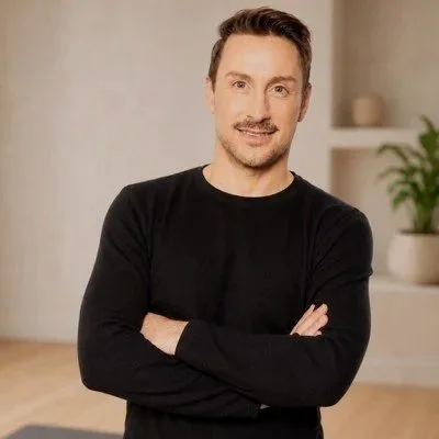 A man with short dark hair and a mustache, wearing a black long-sleeve shirt, standing indoors with arms crossed, smiling.