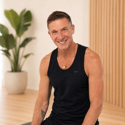 A middle-aged man smiling, wearing a black sports tank top, sitting indoors with a potted plant and wooden panel wall in the background.