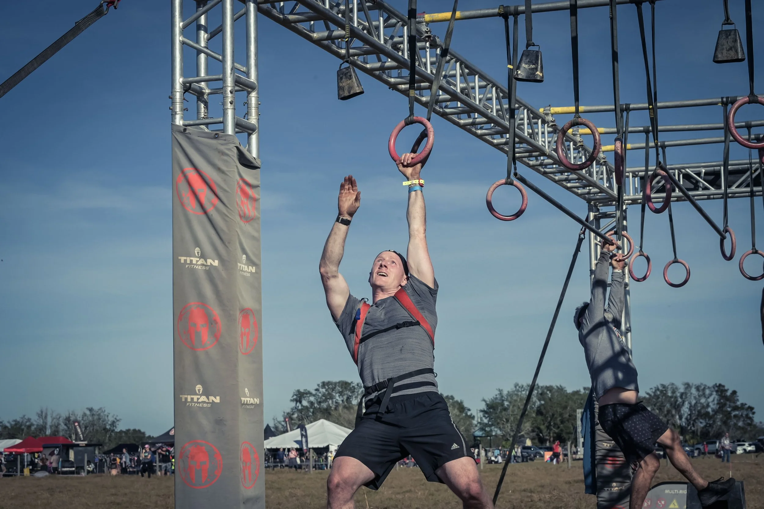 A man participating in an obstacle course event, hanging from gymnastic rings suspended from a metal structure outdoors during daytime, with tents and spectators in the background.