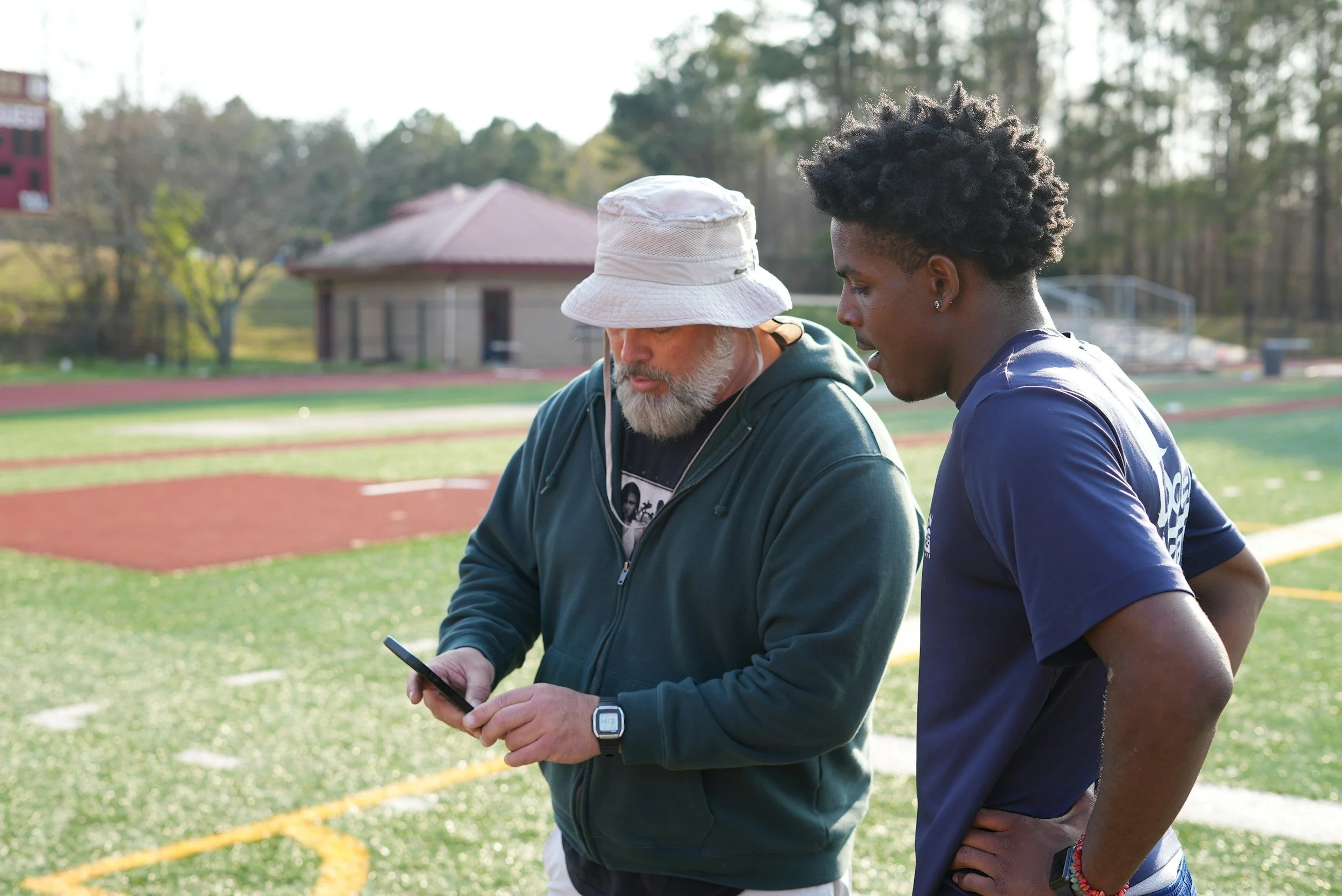 Two men standing on a sports field, one with a white hat and beard looking at a smartphone, the other with curly hair wearing a navy shirt looking at the phone, trees and field background.